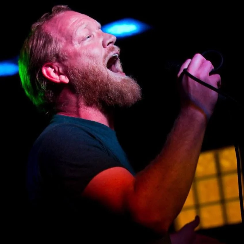 A man with a beard singing passionately into a microphone on stage, with colorful stage lights in the background.