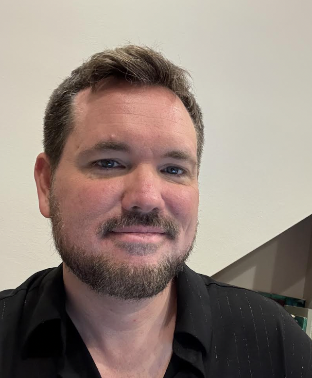 A man with short brown hair, a beard, and blue eyes, smiling, wearing a black shirt, in front of a plain white wall.