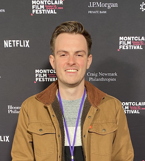 A young man standing in front of a black backdrop with logos and text for the Montclair Film Festival, Netflix, J.P. Morgan Private Bank, and Bloomberg Philanthropies. He is smiling, wearing a tan jacket over a gray shirt, with a purple lanyard around his neck.