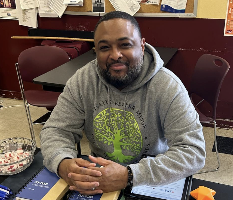 A man with a beard wearing a gray hoodie with a green tree logo, sitting at a desk with notebooks, a bowl of candies, and office supplies.