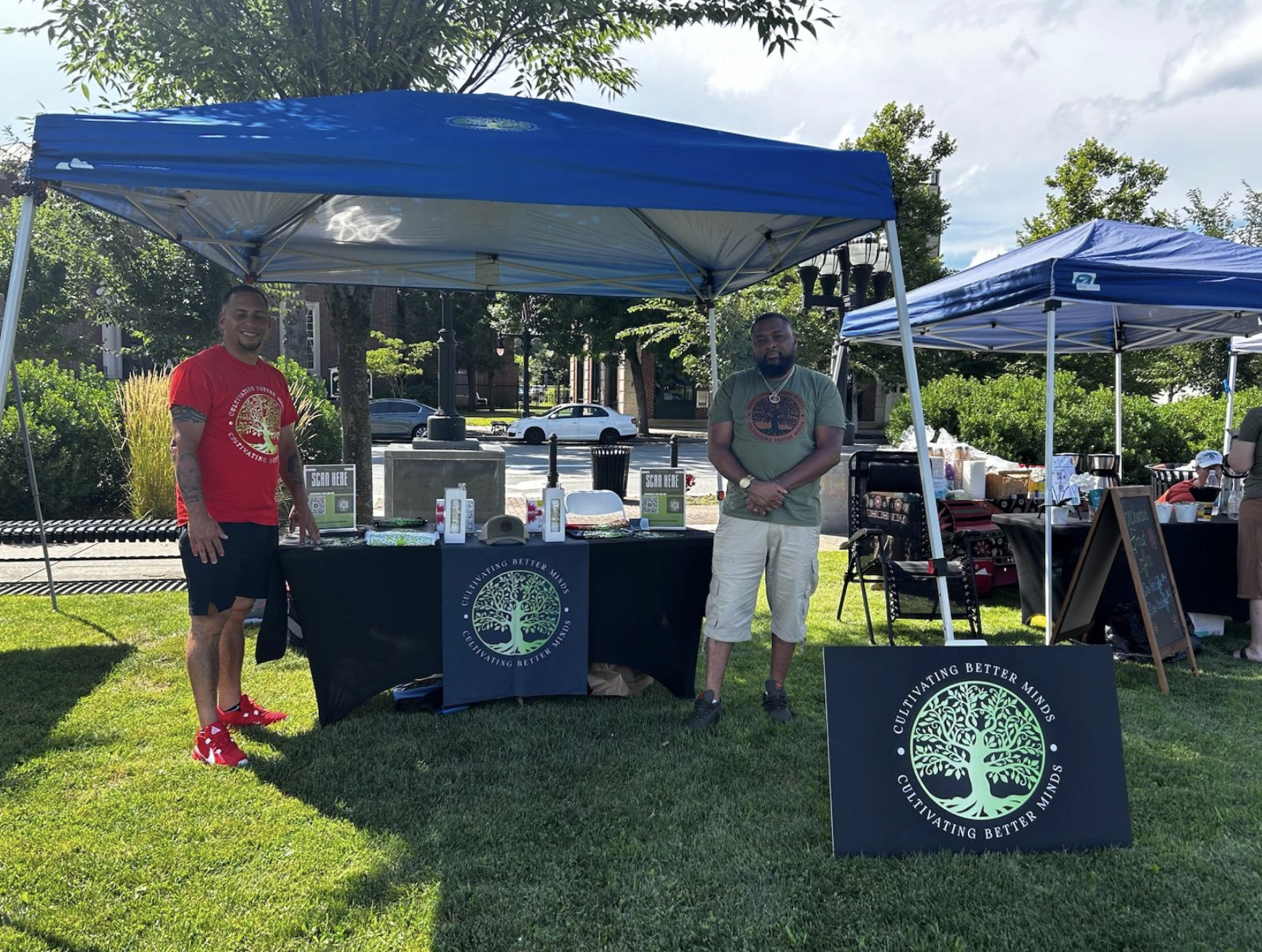 Two men standing at an outdoor booth with a sign that reads "Cultivating Better Minds" and a tree logo, at a park or outdoor event. There are additional tents in the background and people sitting nearby.