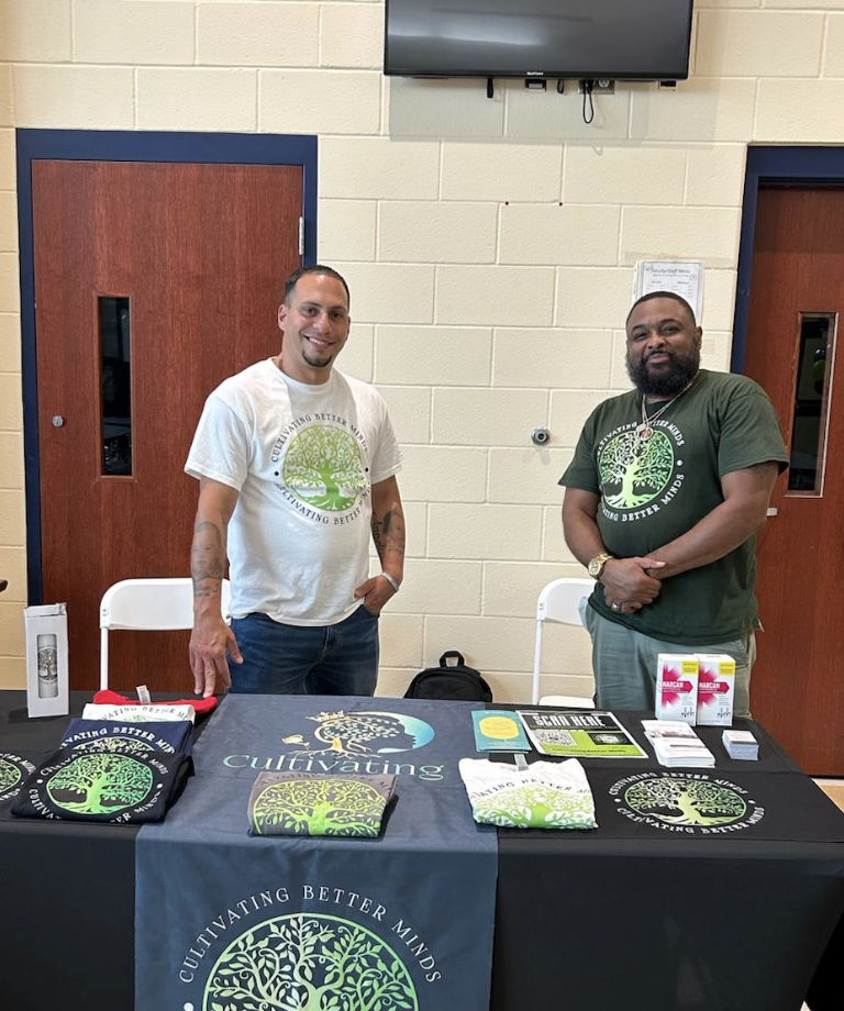 Two men standing behind a table with promotional items including T-shirts, notebooks, and pamphlets, at an indoor event promoting cultivating better minds.
