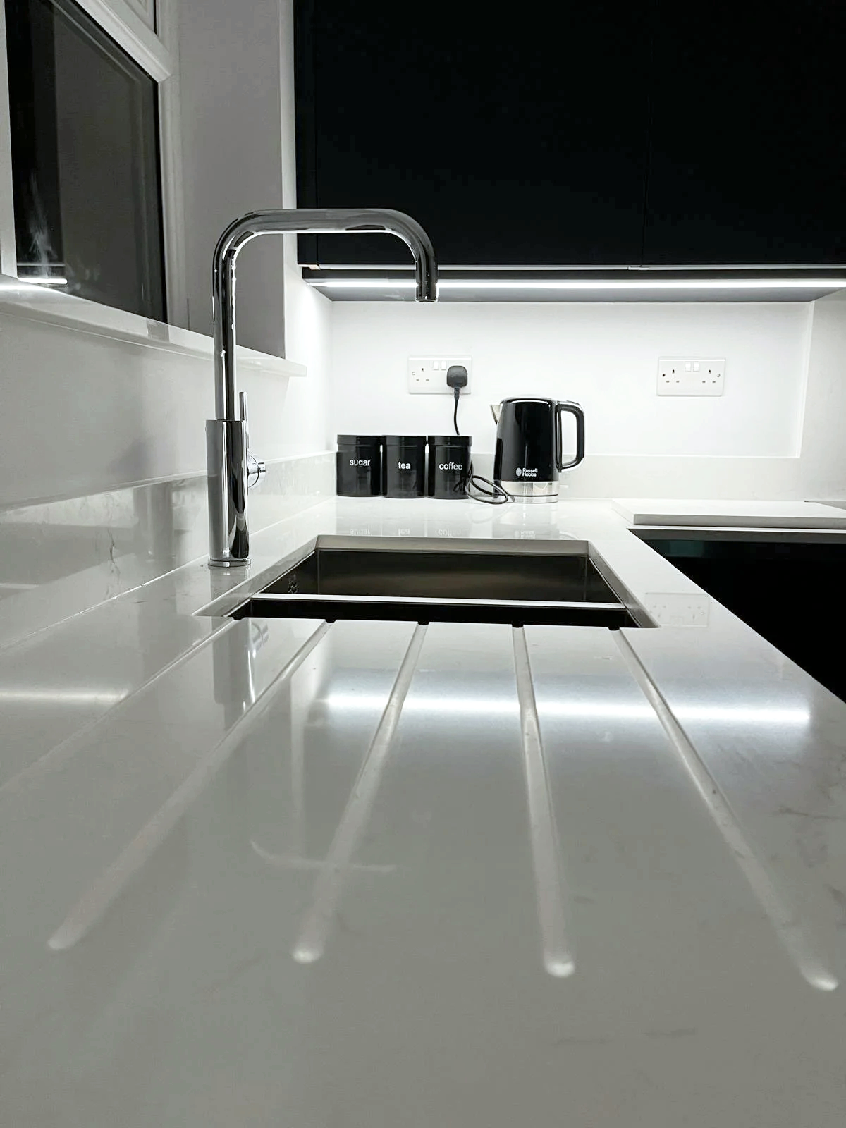 Example of our work - Modern kitchen with white countertops, stainless steel sink, black cupboards, and black kitchen appliances, including a kettle and three canisters labeled sugar, tea, and coffee.
