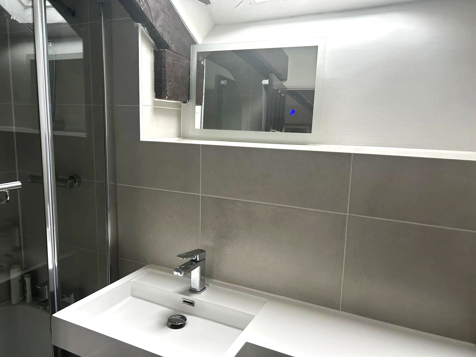 Example of our work - Bathroom with a white sink, grey tiled wall, rectangular mirror, and a shower with glass doors to the left.