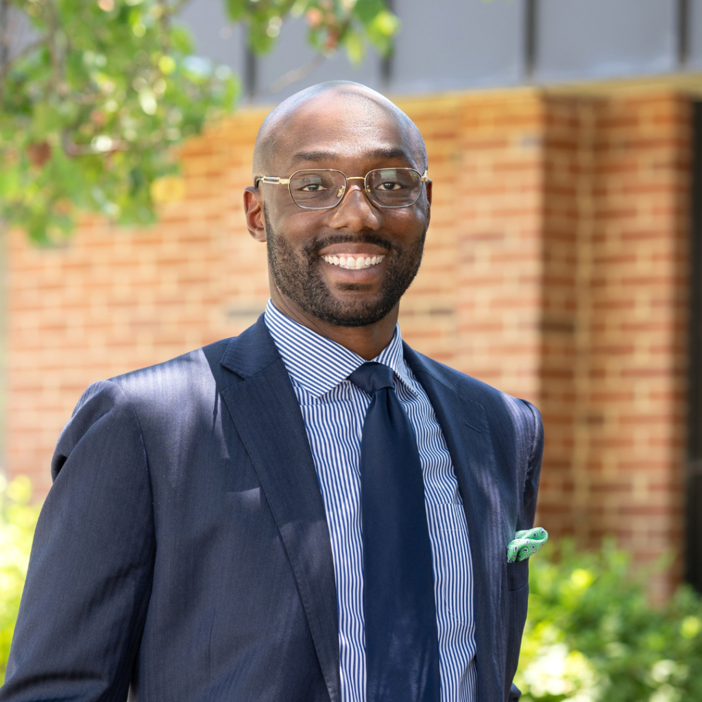 A man in a navy suit, light blue shirt, and navy tie smiling outdoors, with a brick wall and greenery in the background.