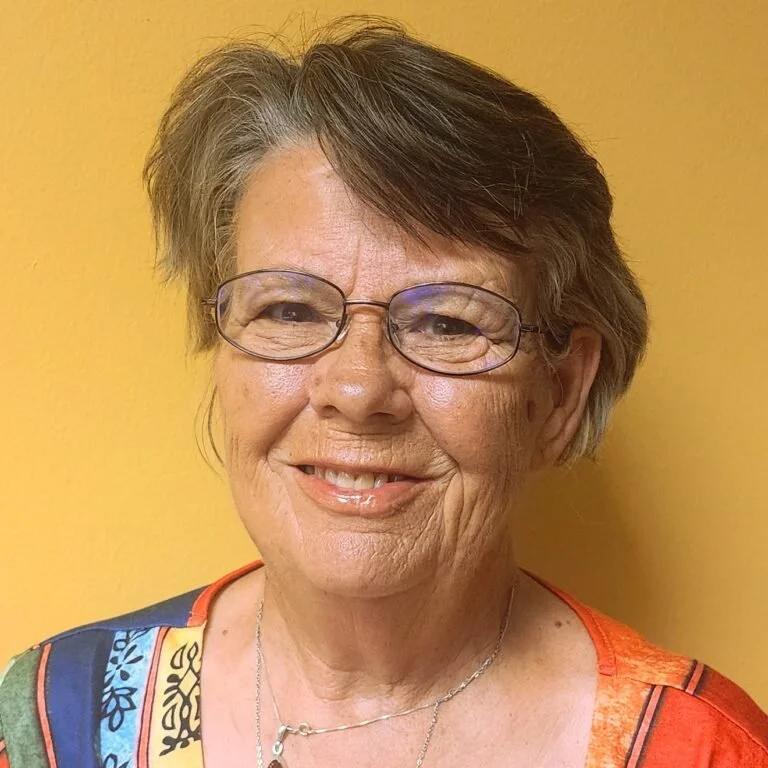 A smiling woman with short brown hair, wearing glasses and a colorful top, standing against a yellow background.