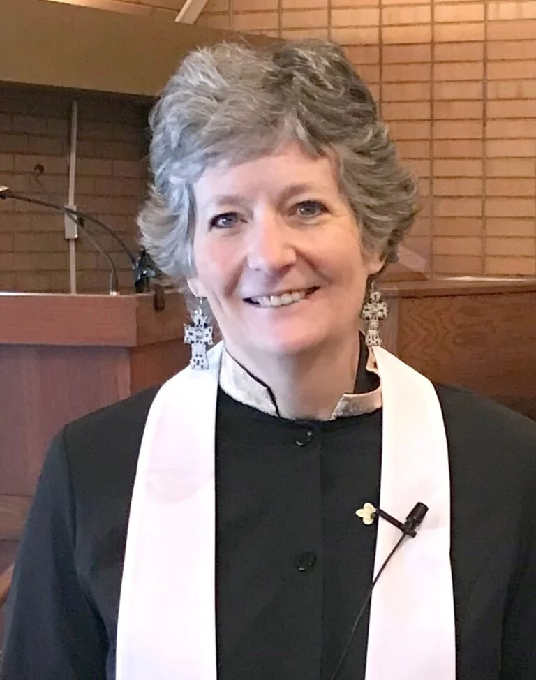 A smiling woman with short gray hair, wearing black clergy attire with white stole, earrings, and a lapel pin, standing inside a church with brick walls.