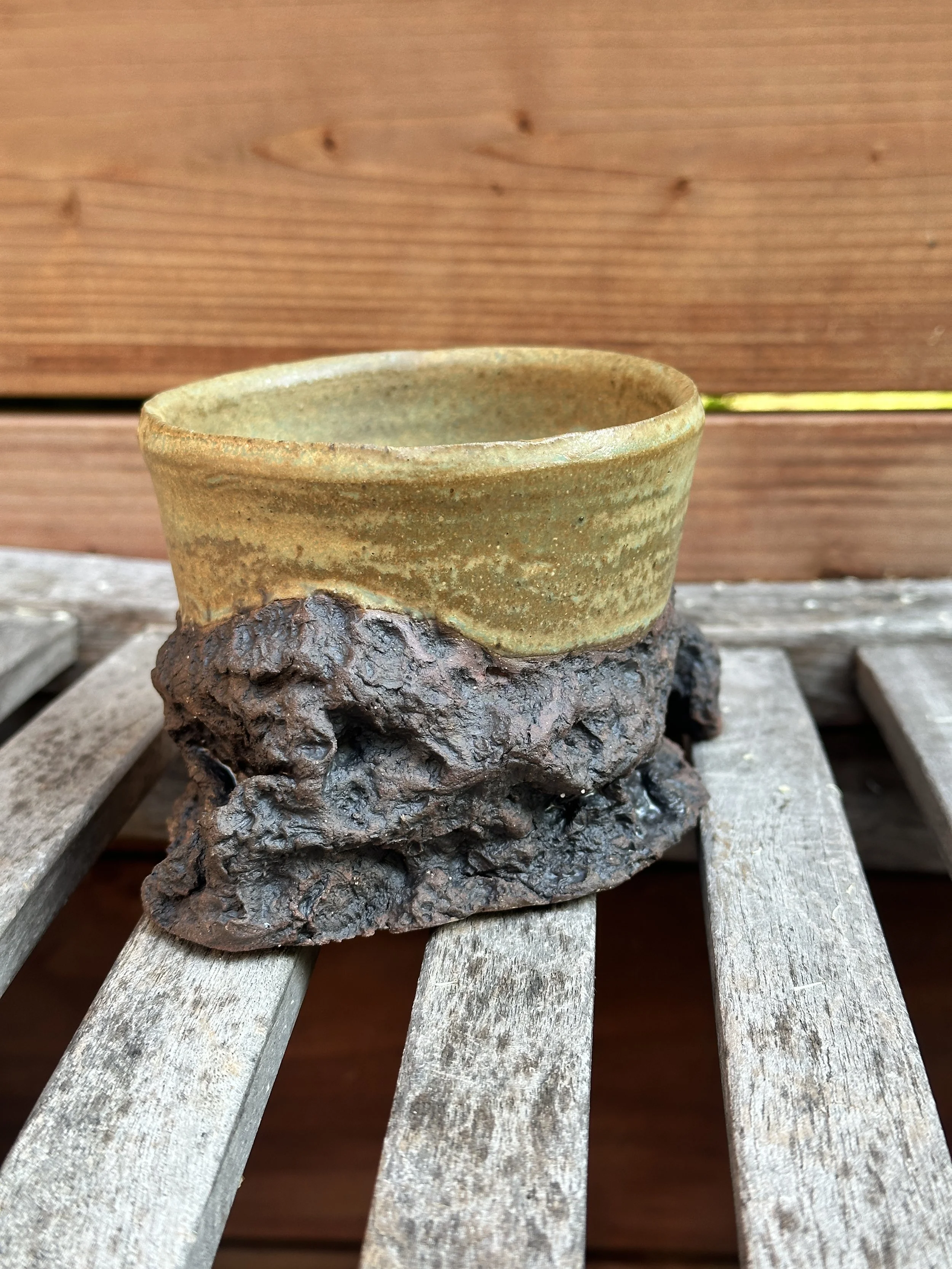 Ceramic mug with a textured, earth-toned glaze sitting on a rough, dark brown base on weathered wooden slats.