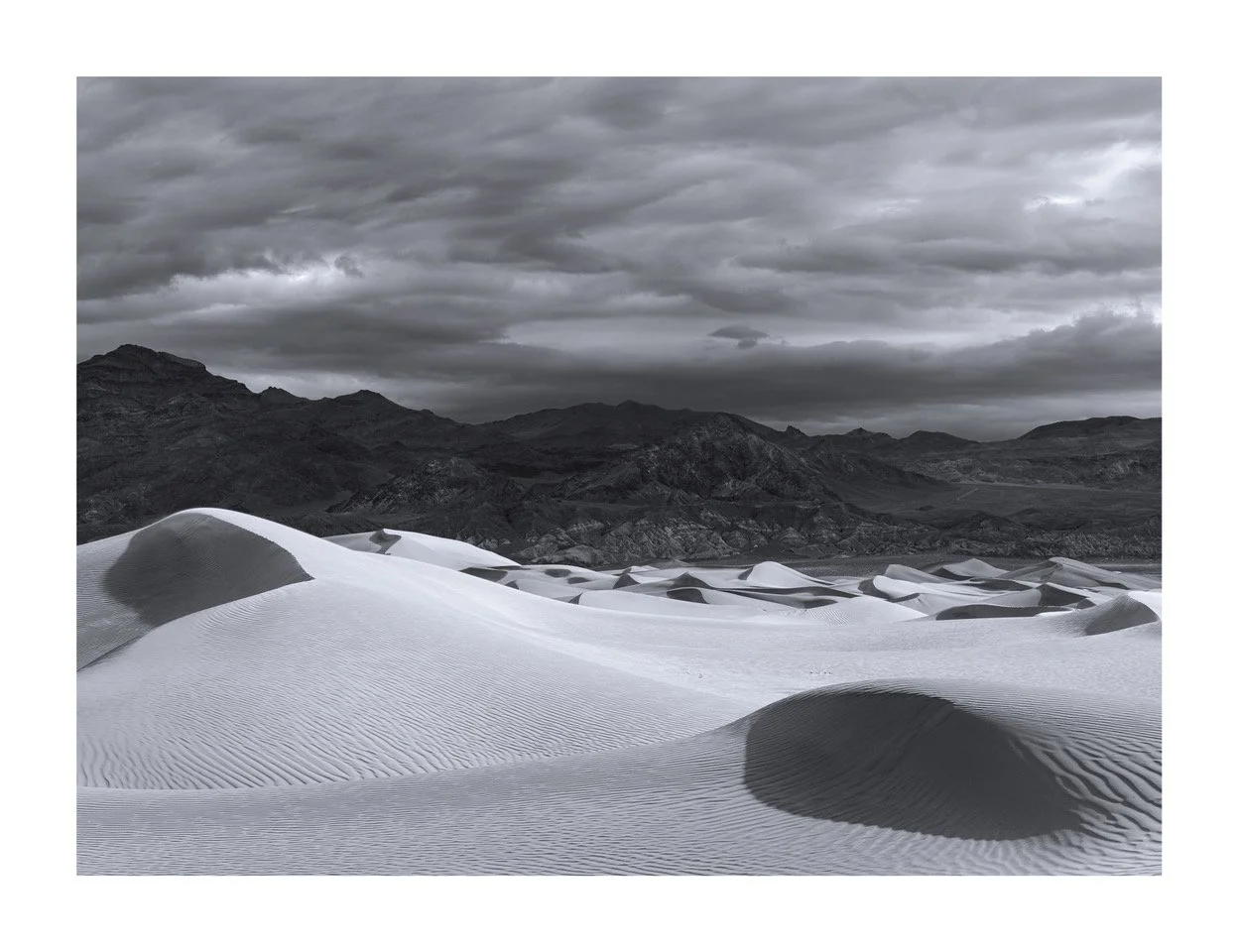 Black and white photograph of sand dunes in a desert with mountains in the background and cloudy sky overhead.