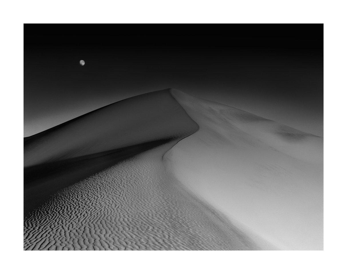 Black and white photo of a sand dune with a moon in the night sky.