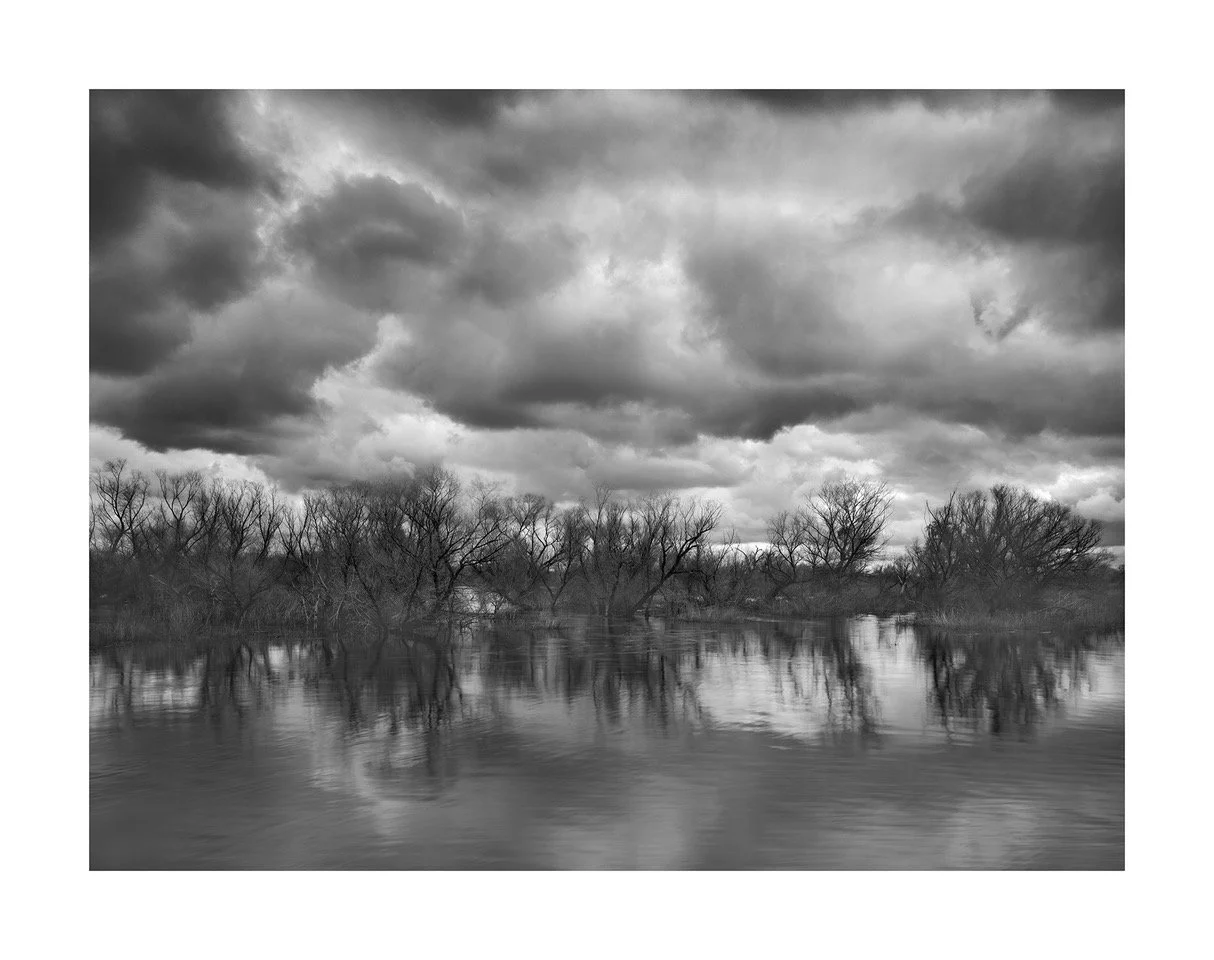Black and white photo of a river with leafless trees along the shoreline and dark, cloudy sky reflected in the water.