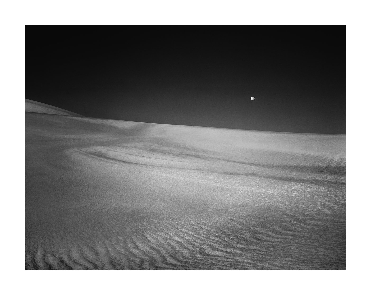 Black and white photo of a desert landscape with sand dunes and a clear, dark sky with a visible moon.