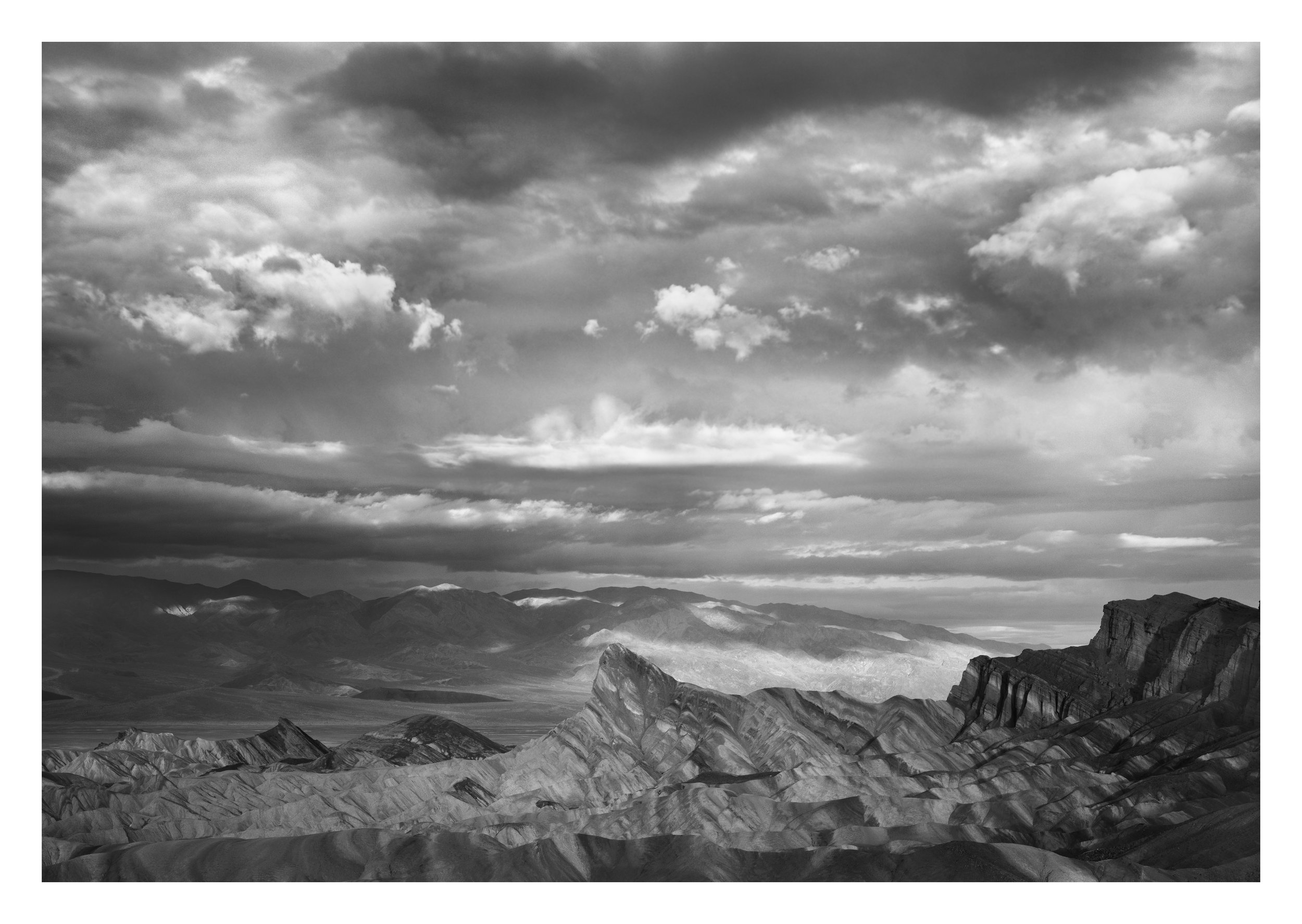 Black and white landscape photograph of mountain ranges with layered rock formations and a cloudy sky overhead.