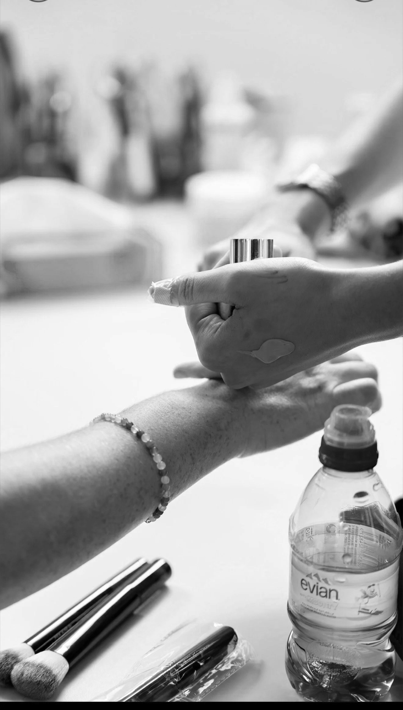Schwarz-Weiß-Foto einer Hand, die eine andere Hand hält. Auf der Haltehand befindet sich ein Armband. Im Hintergrund sind Make-up-Tools und Wasserflasche sichtbar.