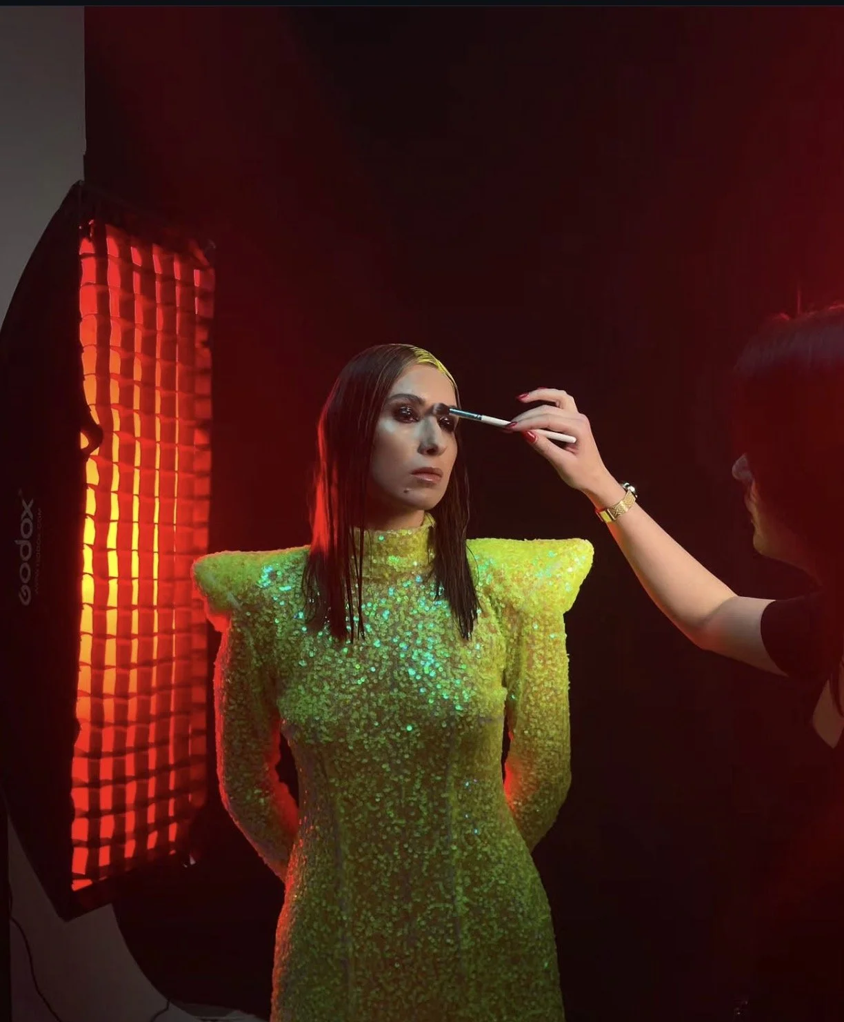 Eine Model wird in einem dunklen Raum mit rotem Licht für ein Fotoshooting vorbereitet, trägt ein grünes, funkelndes Kleid, während eine Person im Hintergrund sie schminkt.