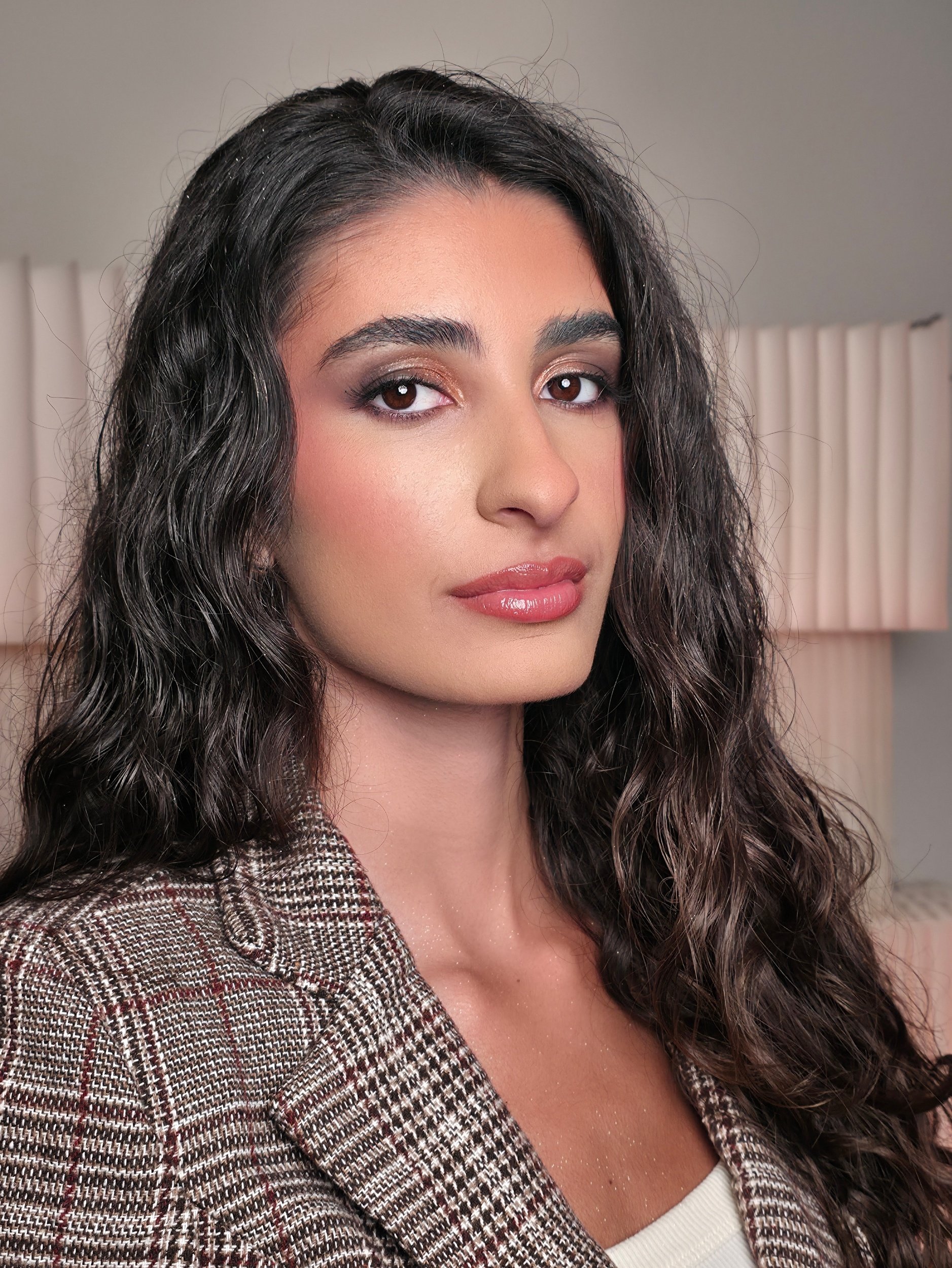 ein Porträt einer jungen Frau mit langen, lockigen dunklen Haaren, dezenten Make-up und einem karierten Blazer, vor einem neutralen Hintergrund.