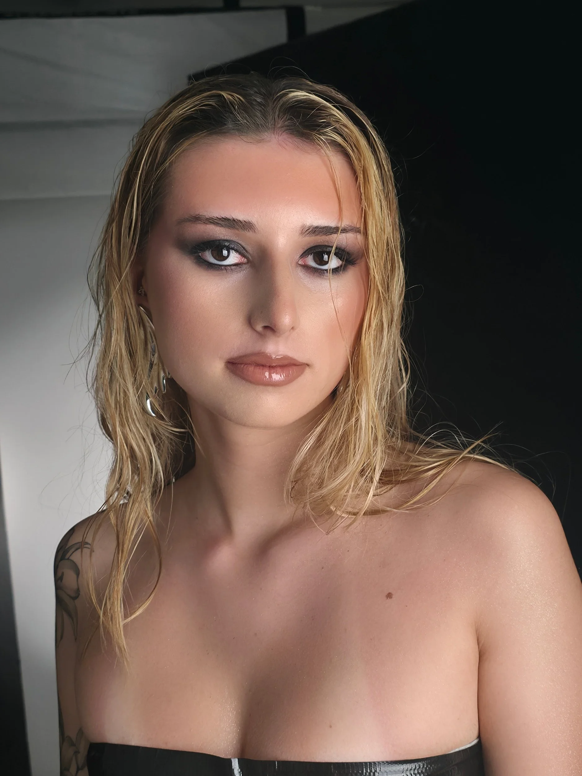 Nahaufnahme eines jungen Mädchens mit nassen, blonden, gewellten Haaren, dunklem Augen-Make-up, unbedecktes Oberteil und Tätowierungen am linken Arm.