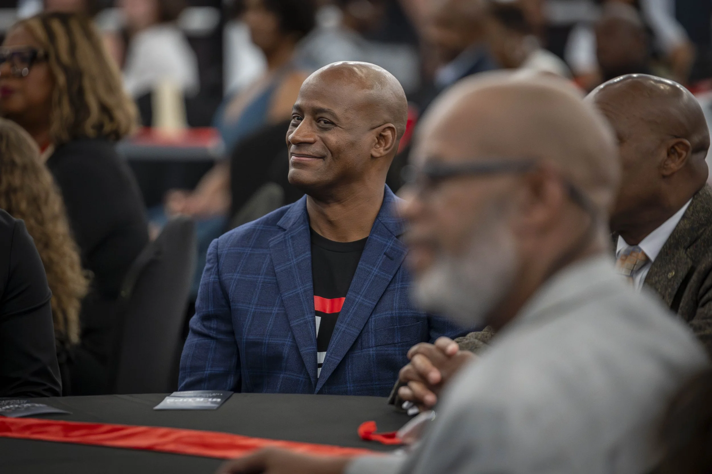 A man with a shaved head wearing a blue plaid blazer is sitting at a table, smiling and engaging with someone across from him. The background shows other people attending an event or conference, with some blurred out.