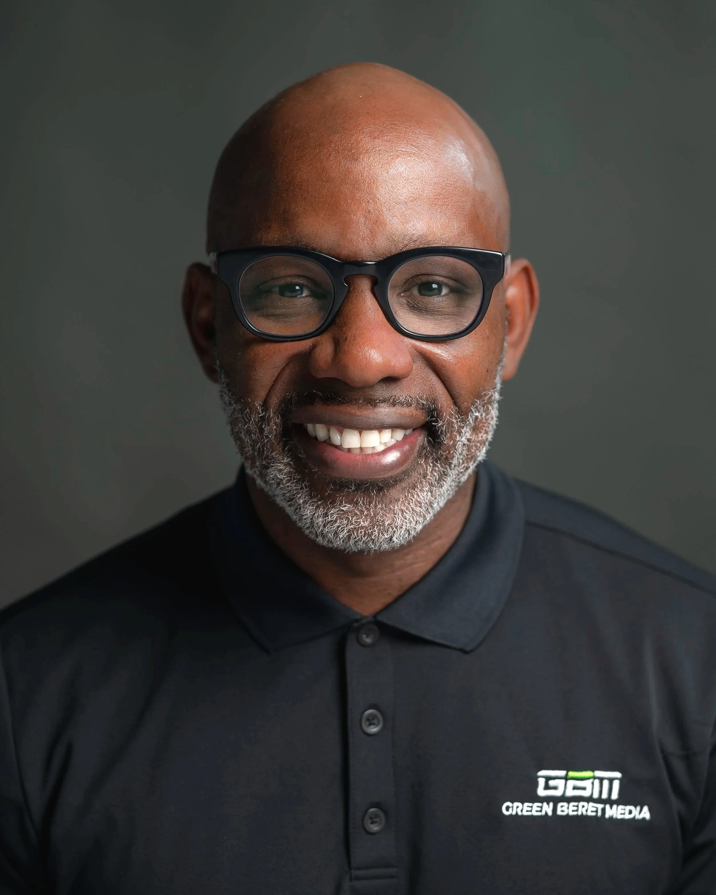 Close-up of a smiling man wearing black glasses and a black shirt with a logo and the text 'GCIM Green Beret Media' on it, against a gray background.