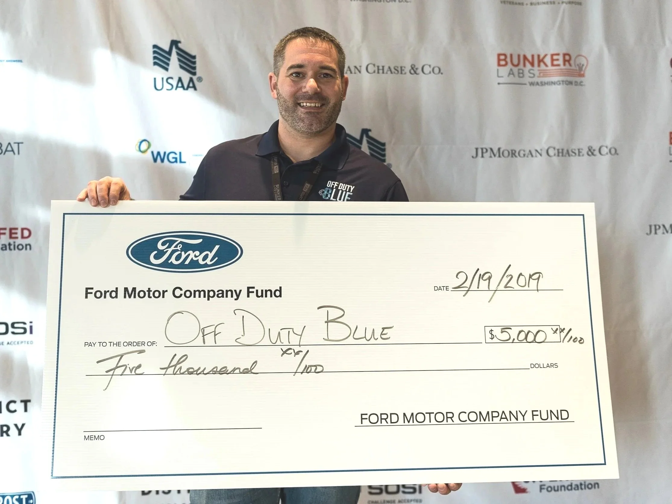 A man smiling and holding a large donation check for five thousand dollars made out to 'Off Duty Blue,' standing in front of a backdrop with logos of USAA and other brands.