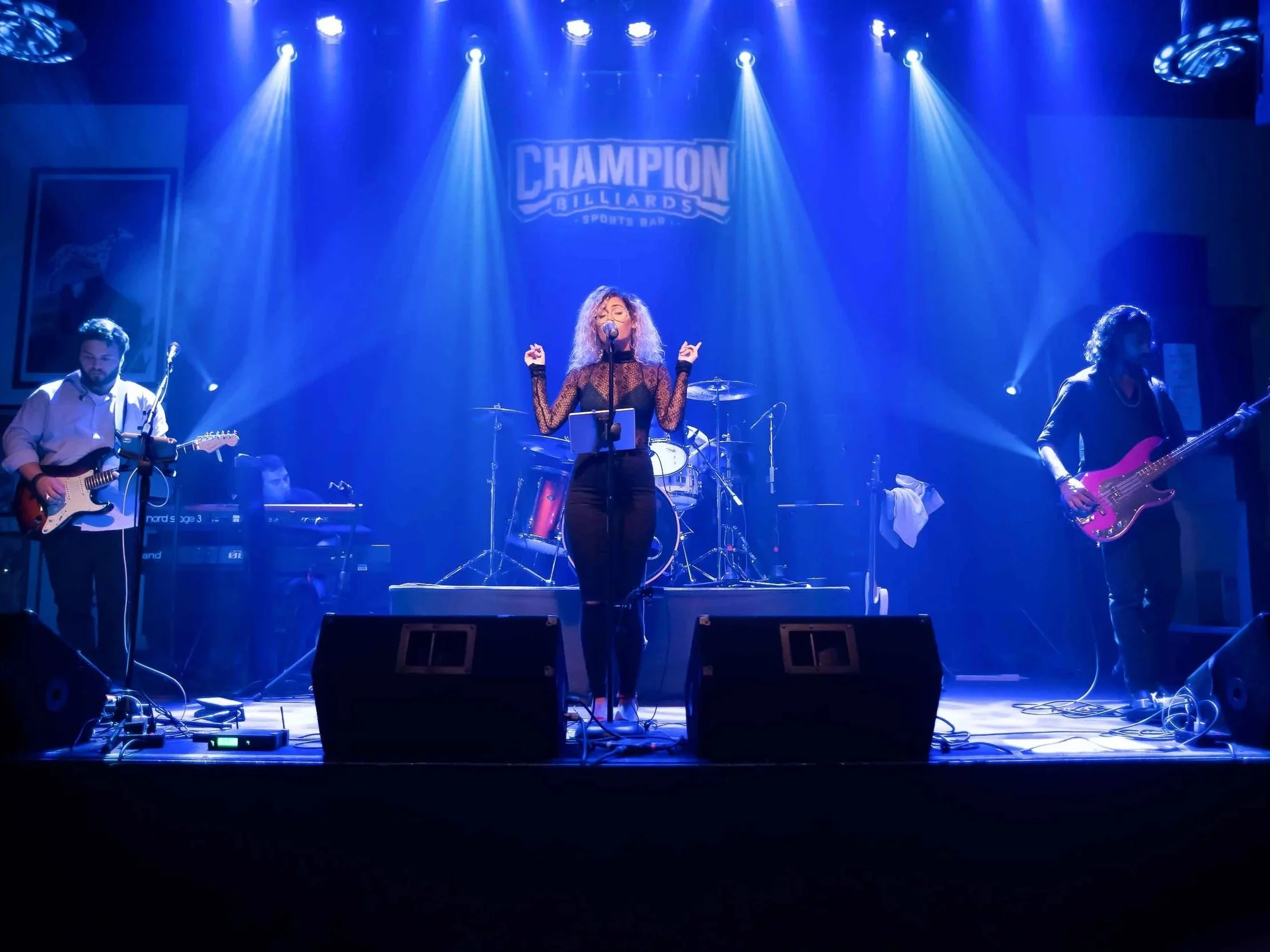 A band performs on stage at Champion Billiards Sports Bar, with three musicians visible under blue stage lighting. The lead singer, a woman with curly hair, stands at the center singing into a microphone. To her left is a guitarist, and to her right, a bassist. The background features the venue's sign and a drum set.