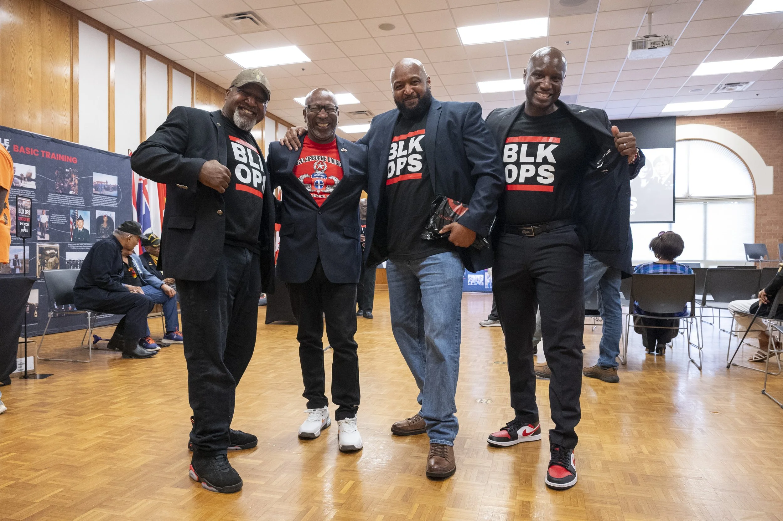 Four men standing together in a room with wooden-paneled walls and a ceiling with fluorescent lights. They are smiling and posing for the photo, wearing casual and semi-formal attire, some with jackets and all with black t-shirts that say 'BLK OPS'.