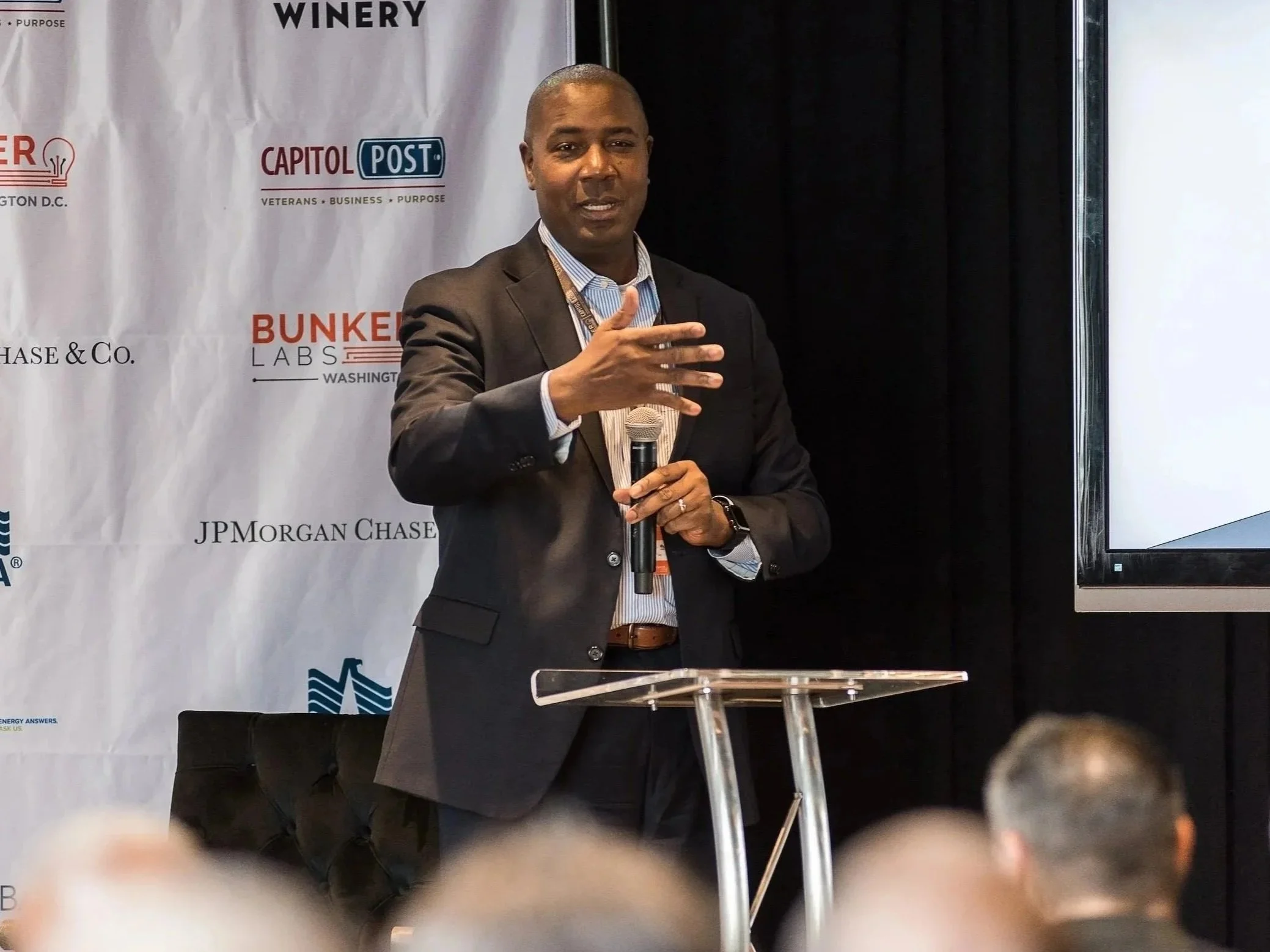 A man in a suit holding a microphone, standing in front of a white background with logos and text related to energy and infrastructure, during a presentation or conference.