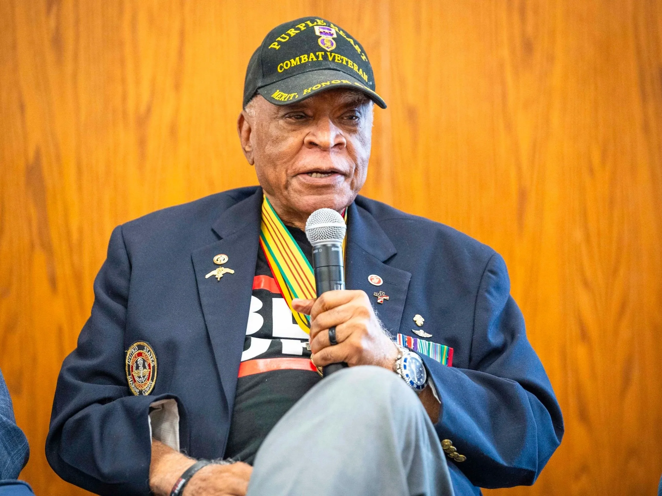 An elderly man wearing a black cap with yellow text reading 'PURPLE HEART' and 'COMBAT VETERAN', dressed in a dark blazer with military medals and insignia, speaking into a microphone in front of a wooden background.