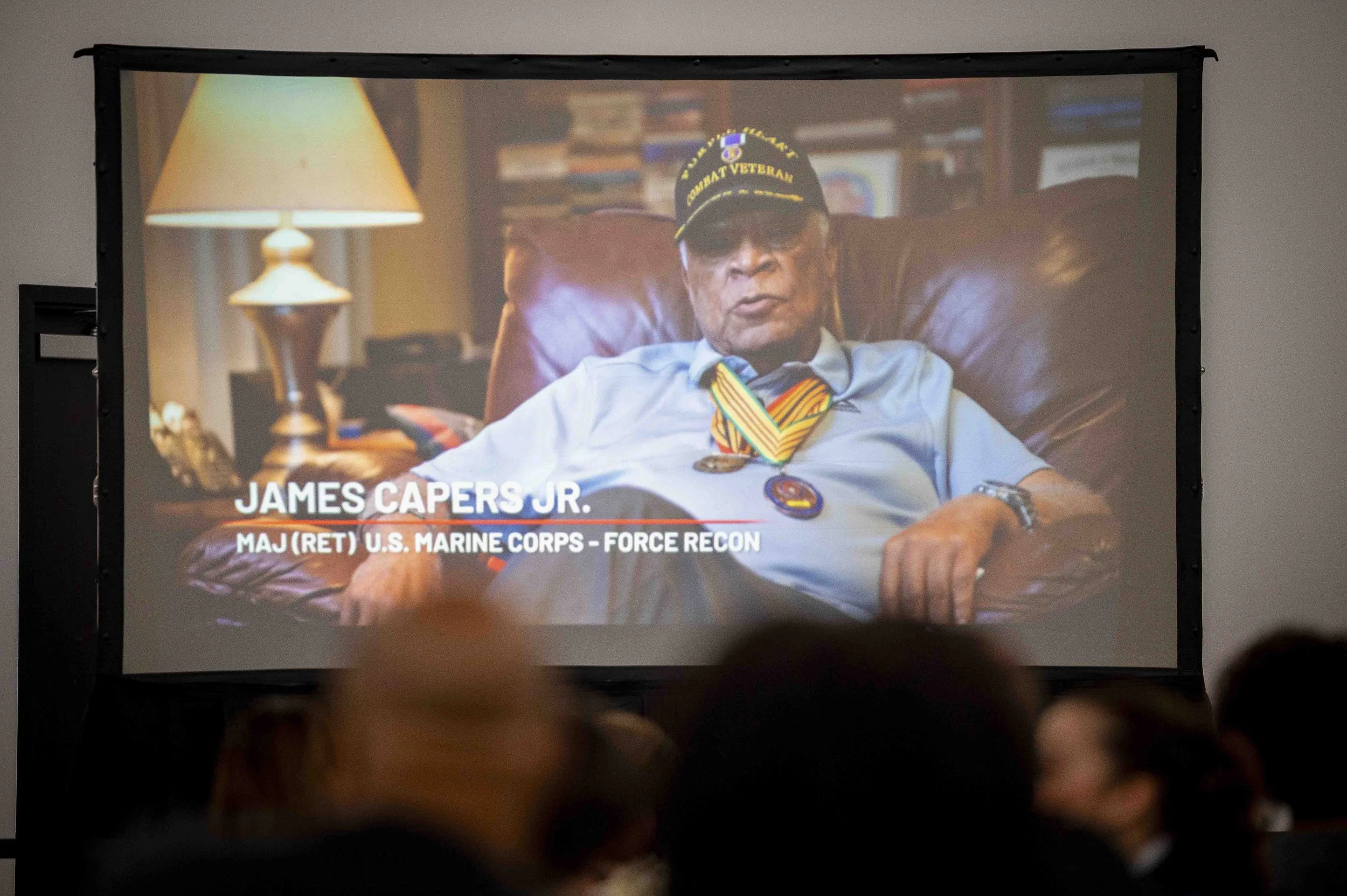 A man wearing a veterans cap and medals, sitting in a leather chair, is featured on a presentation slide. The slide contains text that reads: James Capers Jr., Major (Ret.) U.S. Marine Corps - Force Recon.