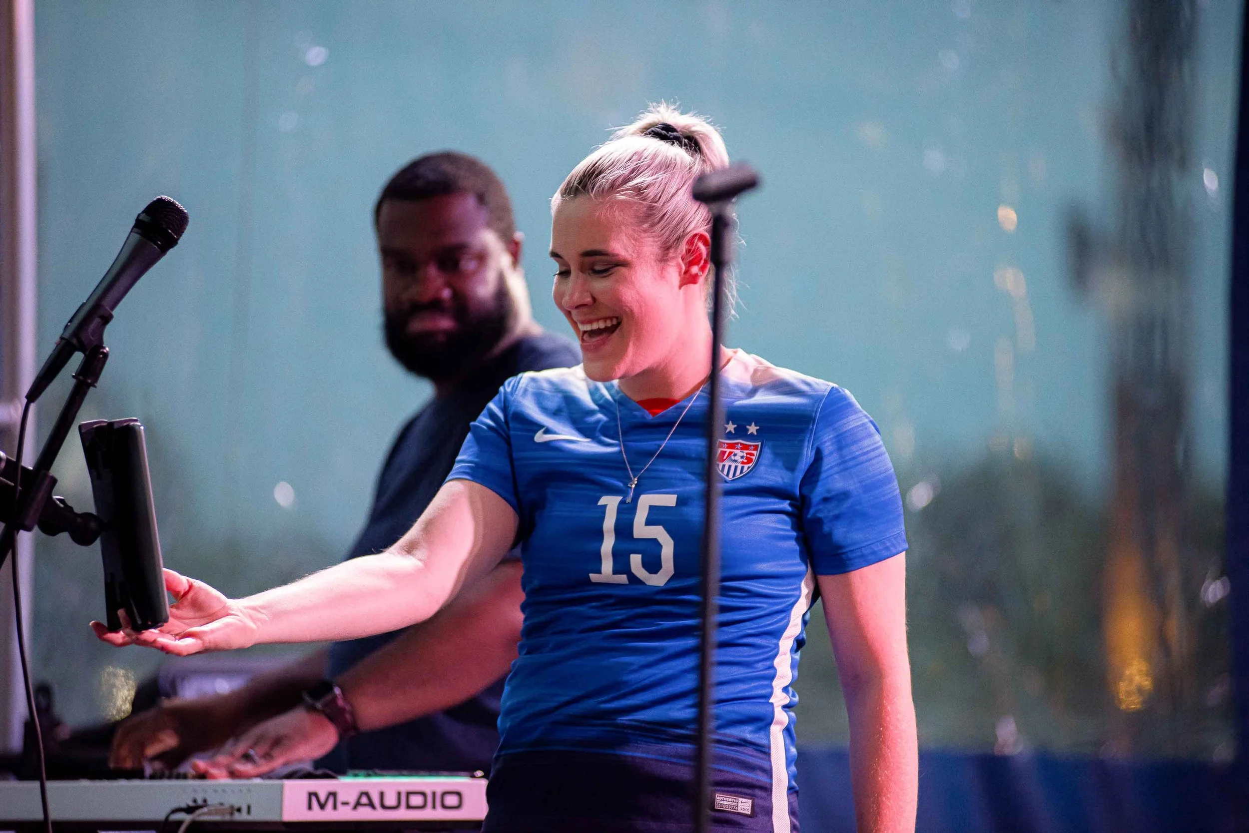 A woman wearing a US women's soccer jersey number 15, smiling and reaching towards a microphone, with a man in the background.