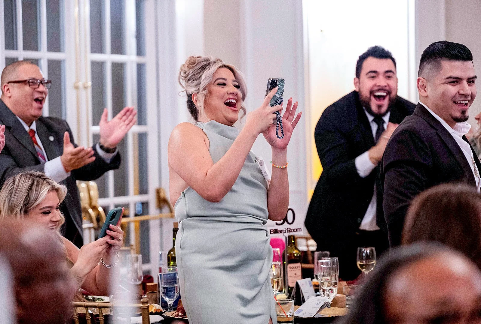 A group of people in formal attire enjoying a lively event, with some taking photos and clapping.