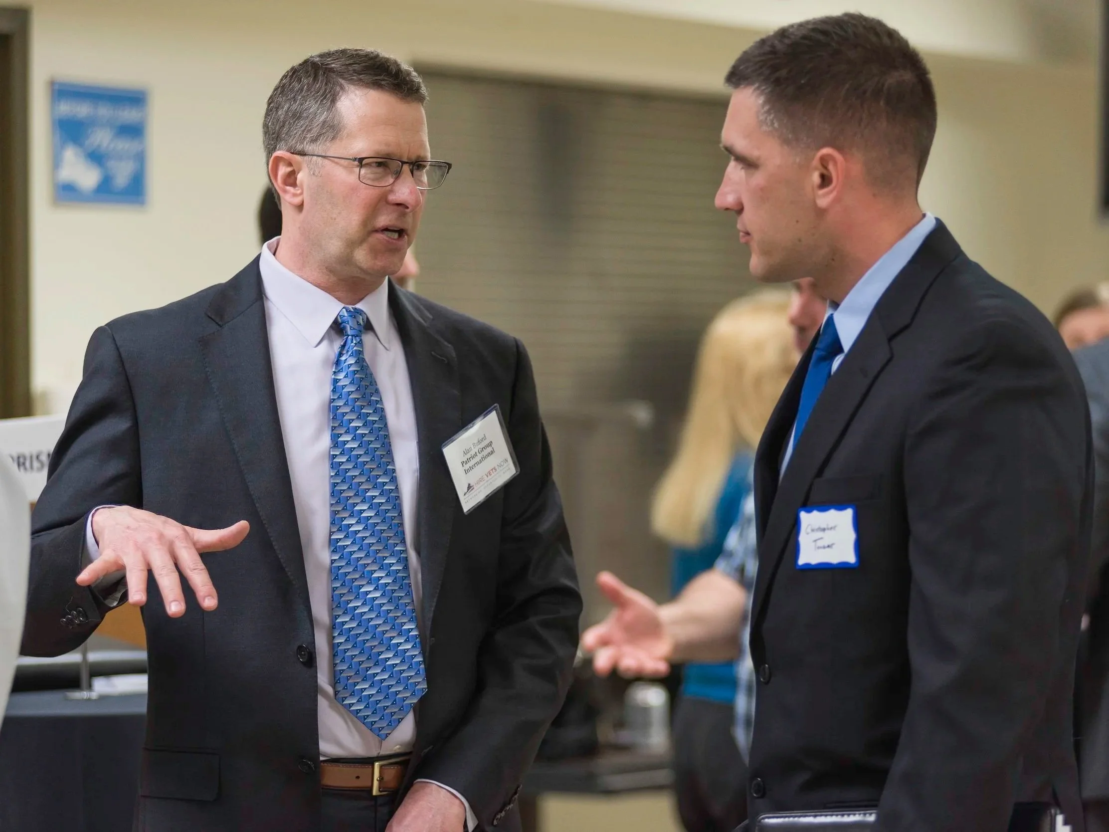 Two men in suits engaged in a conversation at a professional event. The man on the left is wearing glasses and a patterned blue tie, and the man on the right has a short haircut and is wearing a blue tie. Both are facing each other in a lively discussion, with badges on their suits.