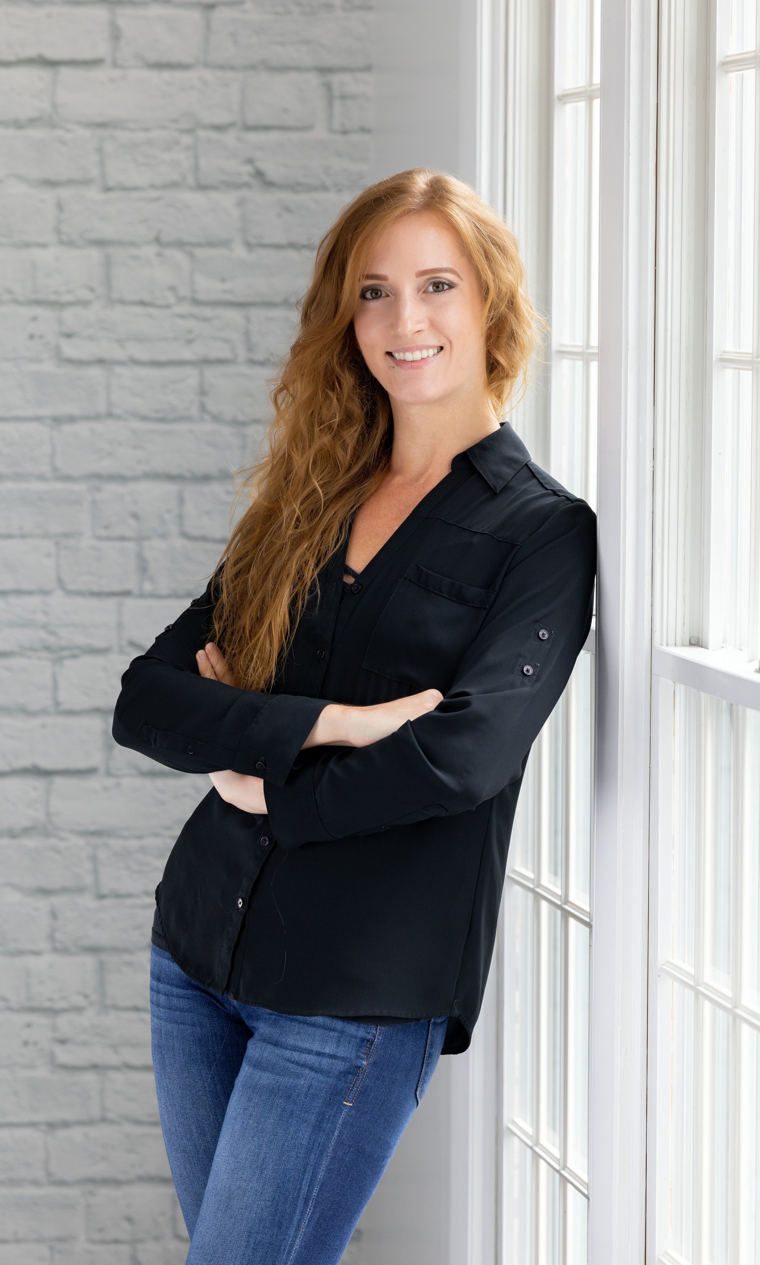 Dr. Kelsey Ramberger PhD with long hair in black shirt and blue jeans standing by window smiling