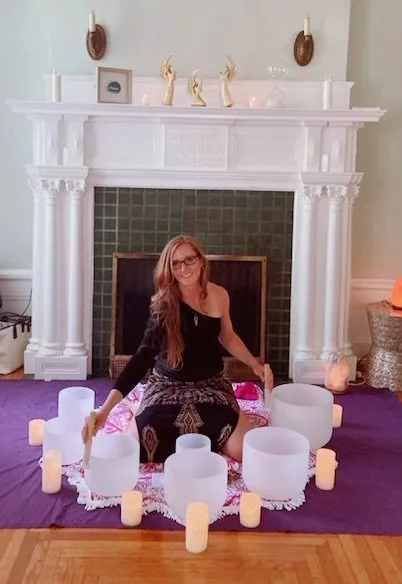 Dr. Kelsey Ramberger, PhD sitting on a purple yoga mat in front of a fireplace, surrounded by white candles and clear crystal singing bowls, holding a mallet.