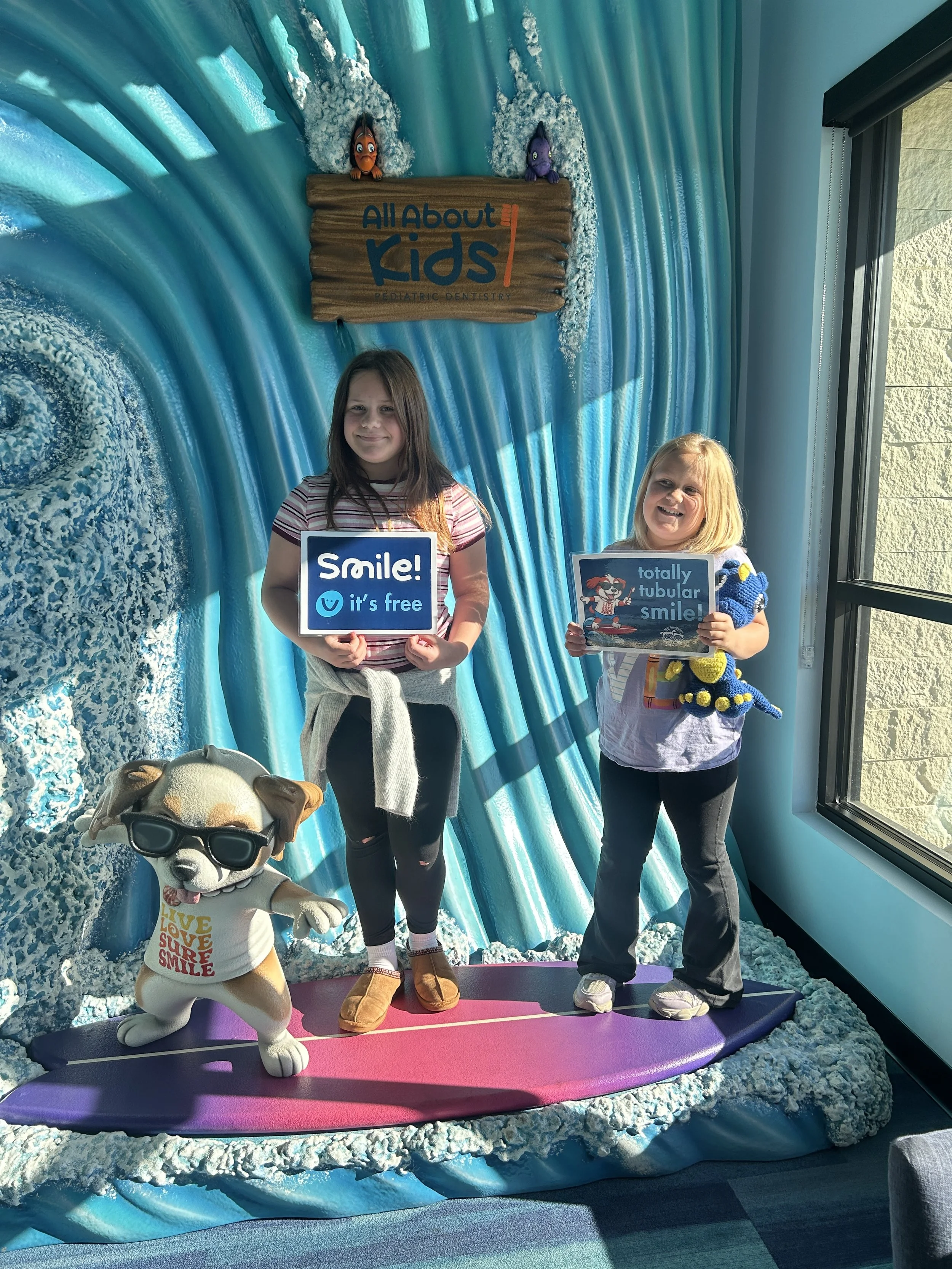 Two young girls standing on a surfboard in a display at a pediatric dentistry office, with sea-themed background. One girl holds a sign that says 'Smile! it's free', the other holds a sign that says 'totally tubular smile'. A plush dog with sunglasses and words 'Love Love Smile' on its shirt is in the foreground. The wall behind features a sign with the text 'All About Kids Pediatric Dentistry' and decorative sea elements.