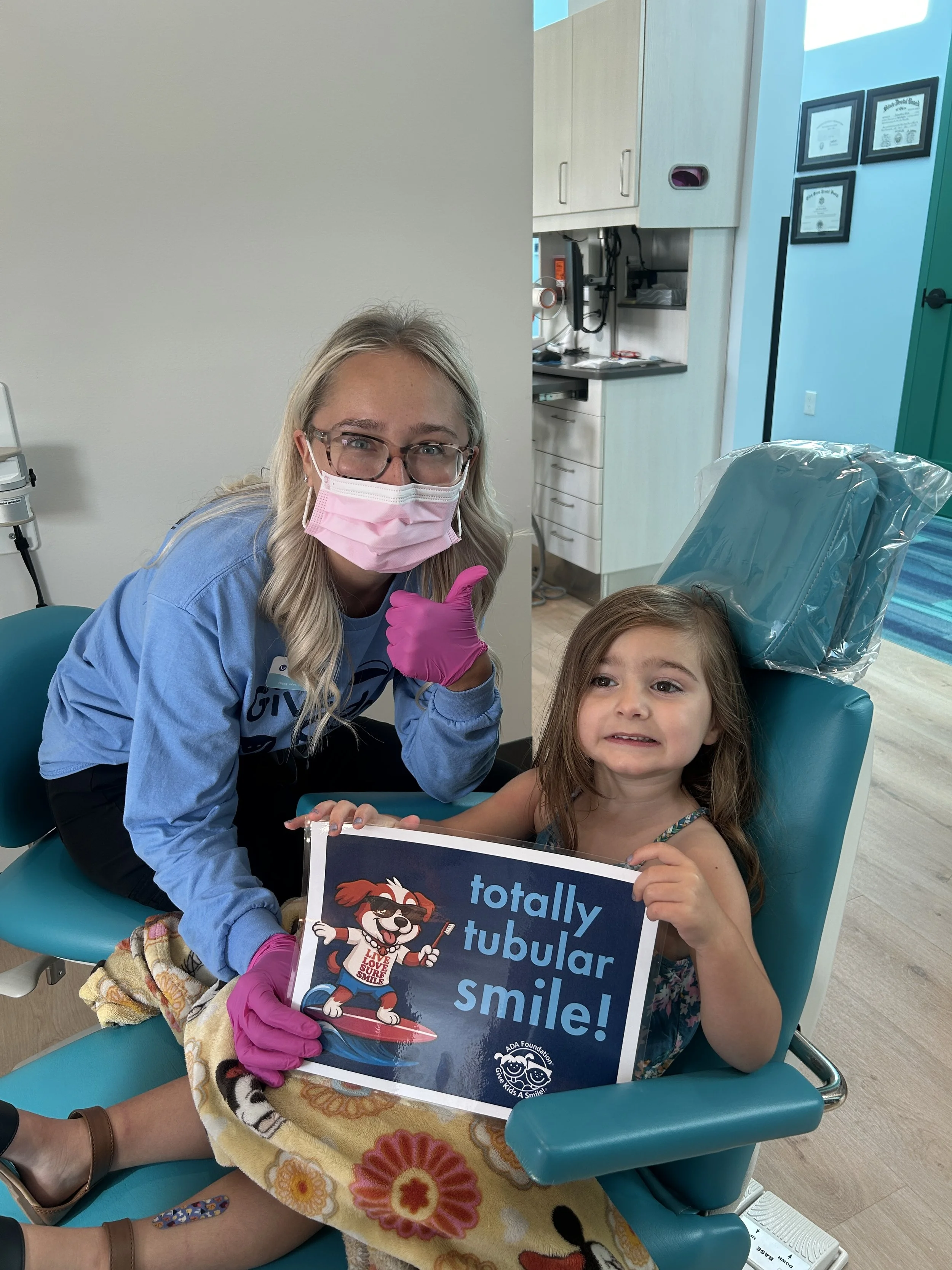 A young girl sitting in a dental chair with a blanket, holding a sign that reads 'totally tubular smile!' with a cartoon dog. A dental professional is beside her giving a thumbs-up, wearing a pink mask and glasses, in a dental office.