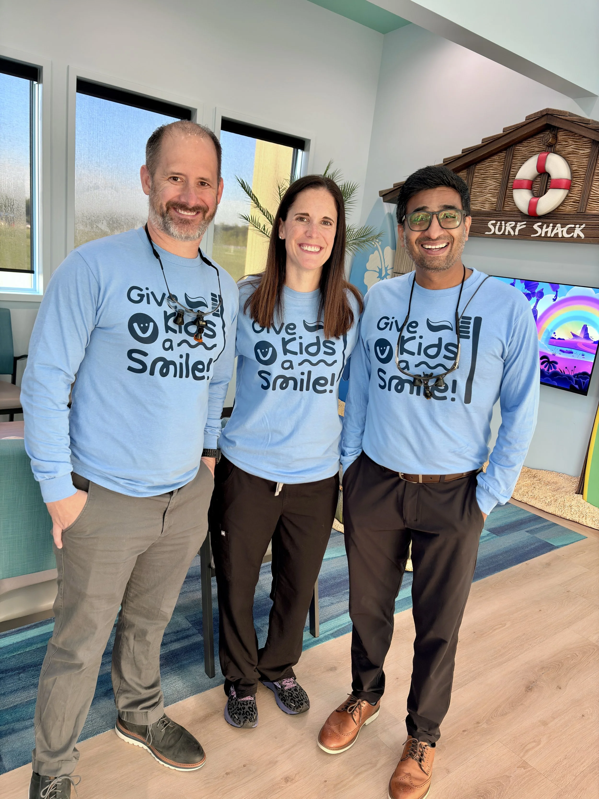 Three smiling adults standing indoors near a surf shack sign, wearing matching light blue shirts with the text 'Give Kids a Smile!'. The person on the left is a white man with a beard, glasses hanging around his neck, and beige pants. The woman in the middle has long brown hair, black pants, and leopard print shoes. The person on the right is a man with glasses and darker skin, wearing dark pants and brown shoes. There is a blue and rainbow-themed wall decoration and a television in the background.