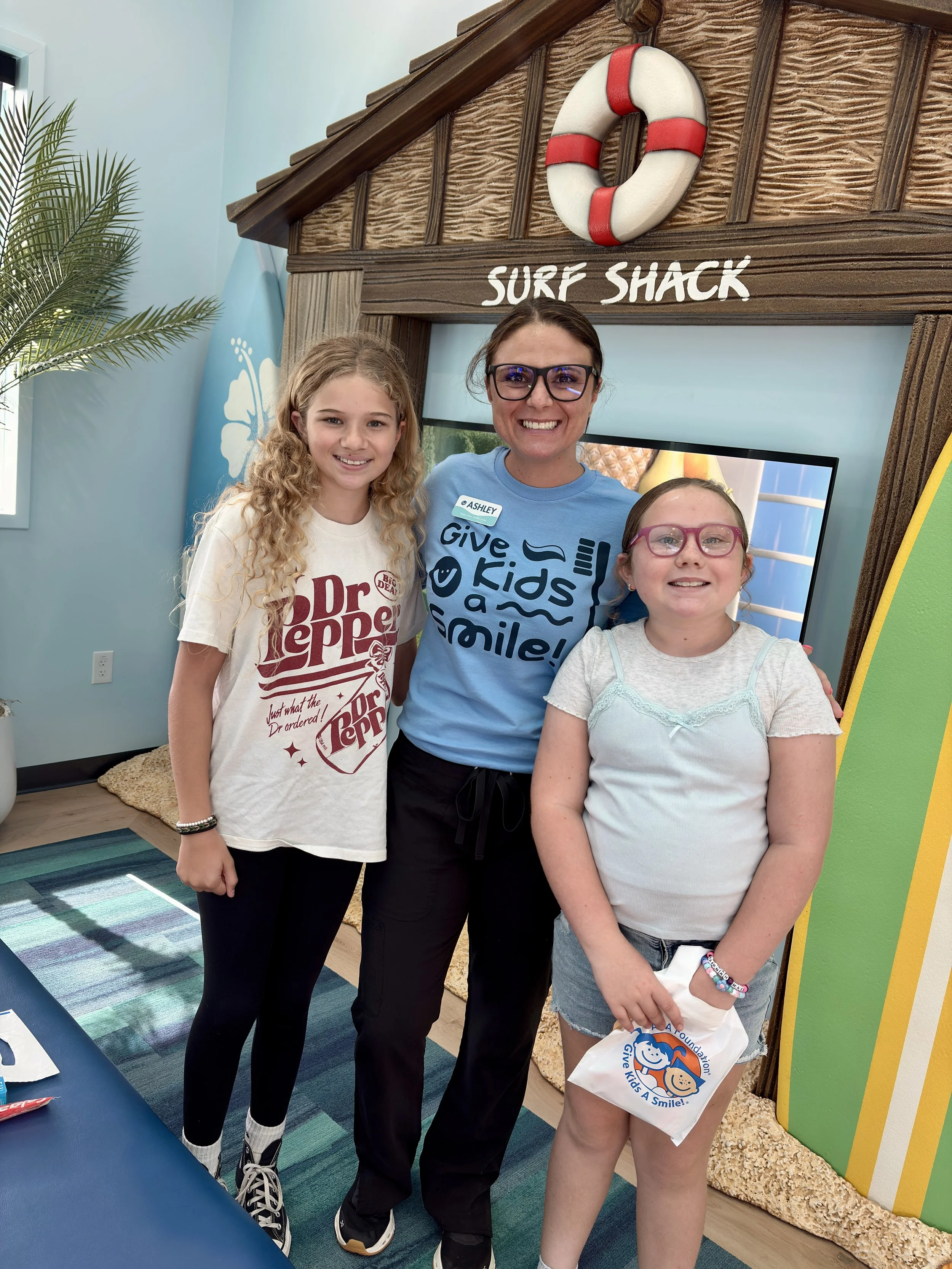 Three people, a young girl, an adult woman, and a young girl, standing together indoors. They are smiling and appear to be at a children's charity event called "Give Kids A Smile" with a surf shack theme. The background shows a television screen, a decorative surfboard, and a lifebuoy on a wooden structure labeled "Surf Shack." The young girl on the left has long curly blonde hair and is wearing a White "Dr. Pepper" T-shirt, black pants, and sneakers. The woman in the middle has brown hair, glasses, and a light blue T-shirt with the words "give kids a smile!" printed on it, and is wearing black pants. The girl on the right has straight brown hair, pink glasses, and a gray T-shirt with lace details, holding a bag with a smiling cartoon face logo.