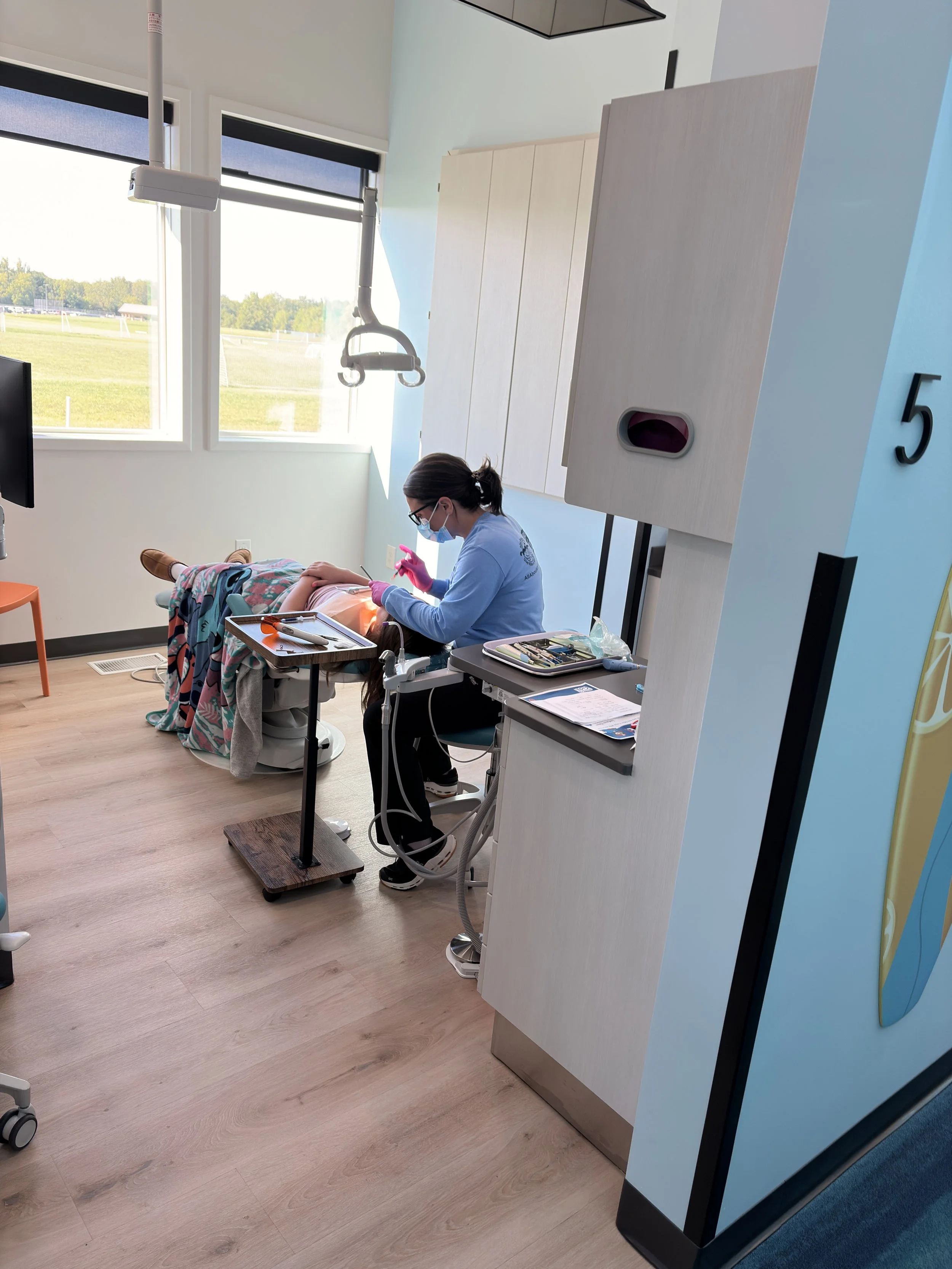 A medical professional wearing a mask and pink gloves preparing a patient for an injection in a hospital room with large windows and a view of grassy fields outside.