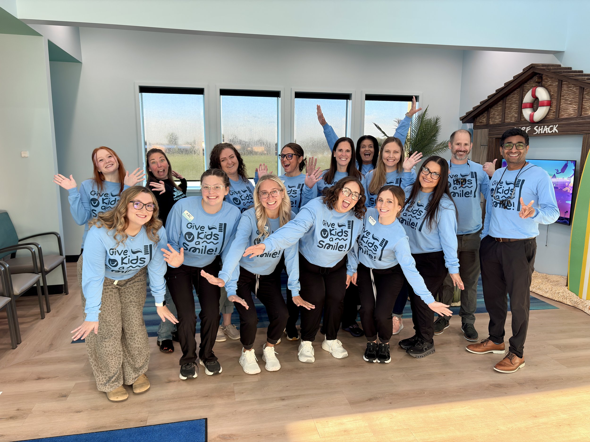 A group of smiling people wearing matching blue T-shirts with the logo 'Give Kids a Smile!' on them, standing in a room with three large windows and a decorative wooden beach shack structure in the background.