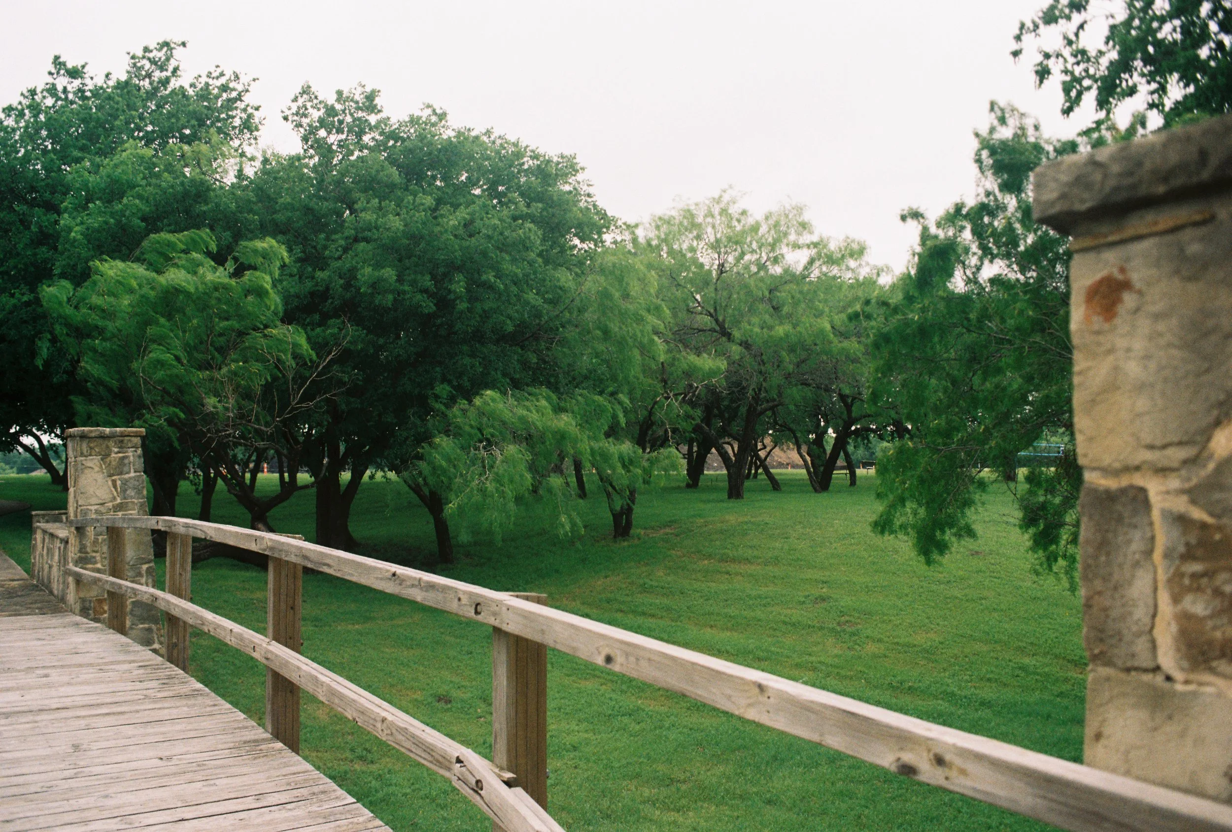 View of a park or garden with lush green trees, grass, and a wooden railing along a pathway, partly framed by a stone post.