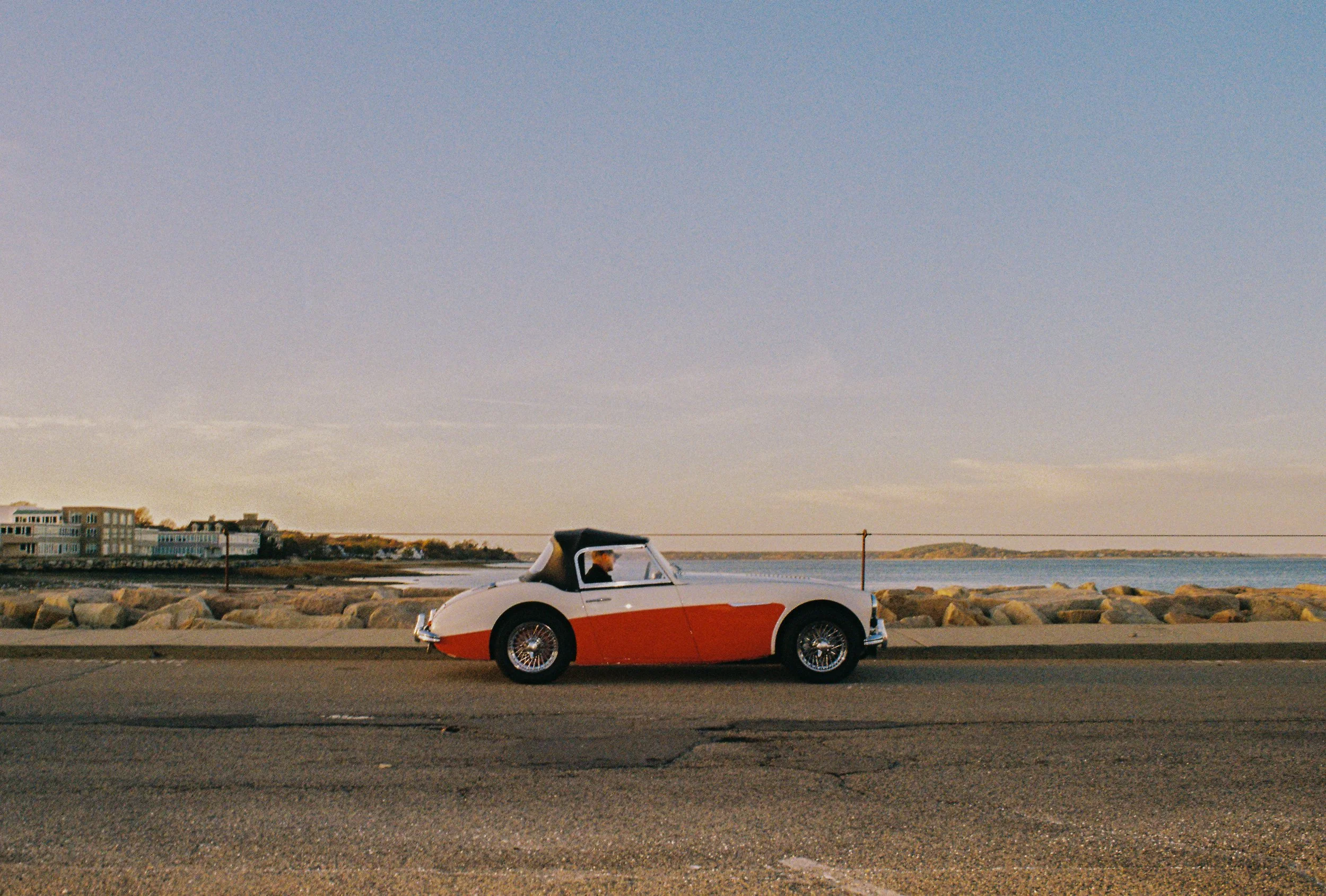 A vintage red and white convertible car parked near a waterfront with rocks and a distant shore in the background, under a clear sky.