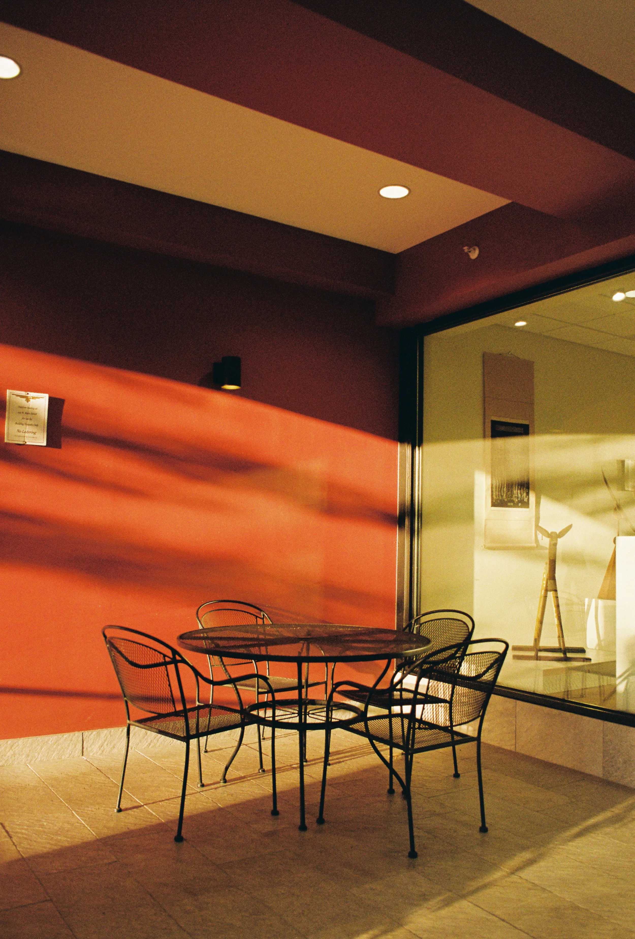 Empty outdoor patio with black metal table and chairs, illuminated by sunlight, with a red wall and a window showing interior decor.
