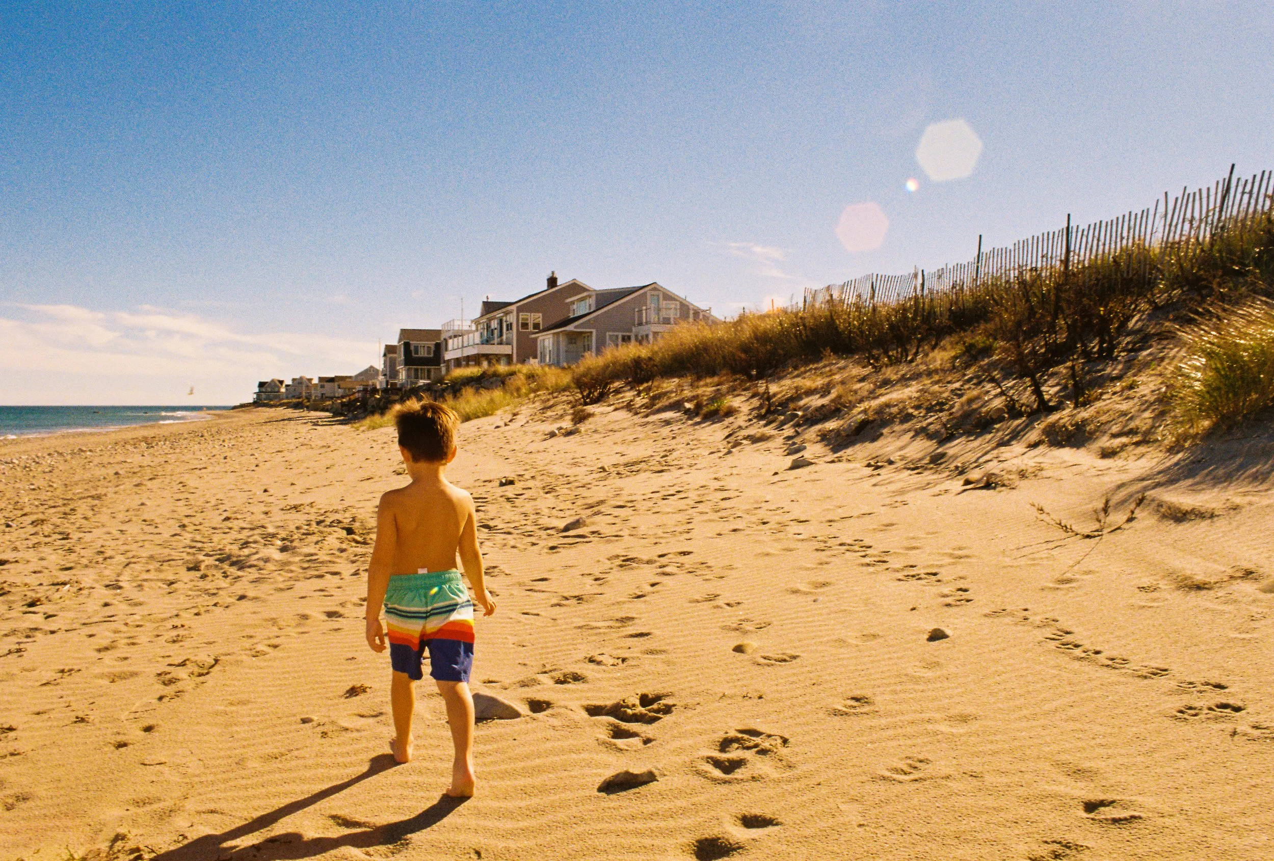 A young boy walking barefoot on a sandy beach towards distant houses, with footprints behind him and a clear blue sky overhead.