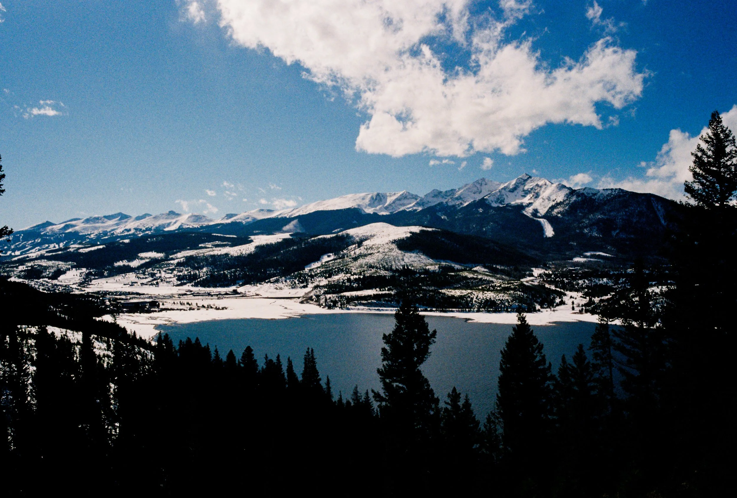 Snow-covered mountains, a lake, and a forest of pine trees under a partly cloudy blue sky.
