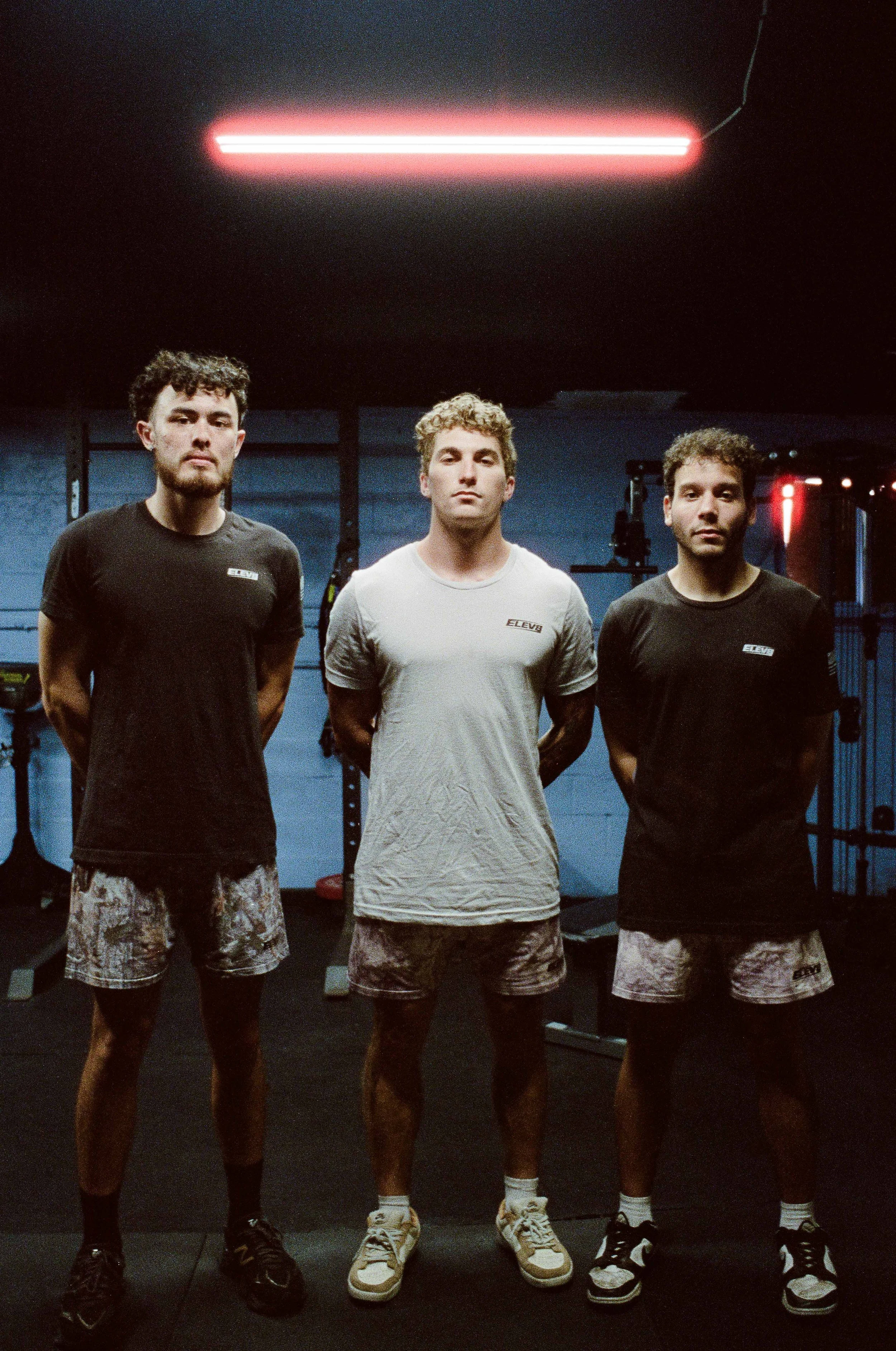 Three men standing side by side in a gym, wearing athletic shirts and shorts, with gym equipment behind them and a neon light overhead.