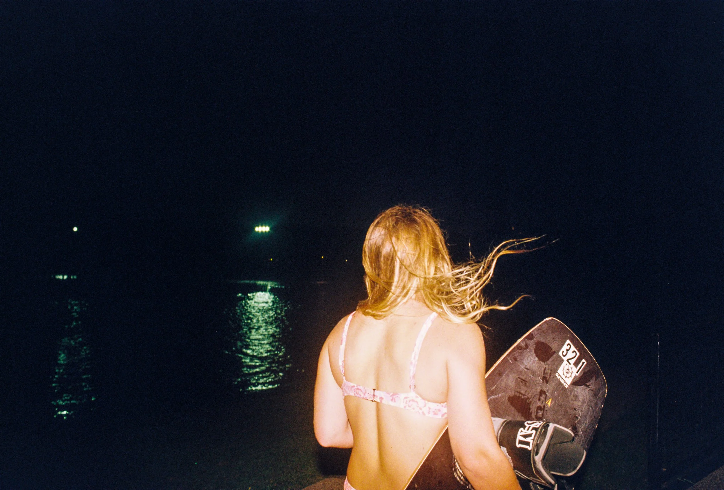 A woman with blonde hair in a swimsuit holding a wakeboard at night, standing near the water with distant reflections of lights.