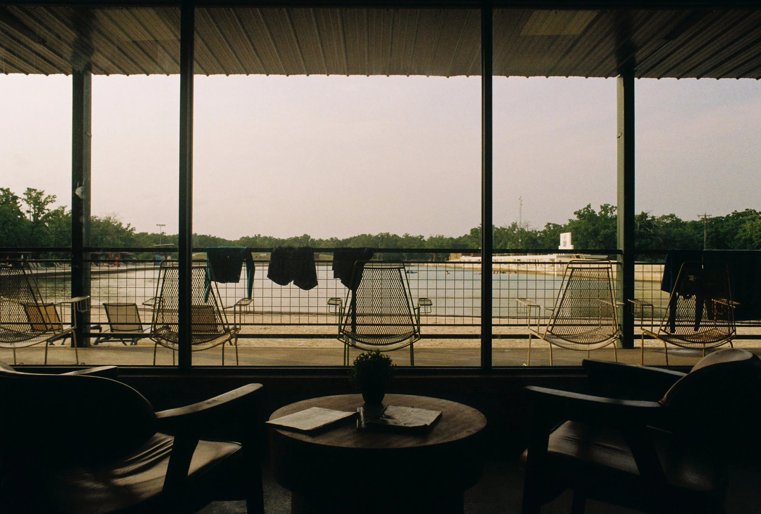 Indoor scene with a circular table, two chairs, and a view of a river through a large window, with outdoor chairs and laundry hanging on a clothesline on a balcony.
