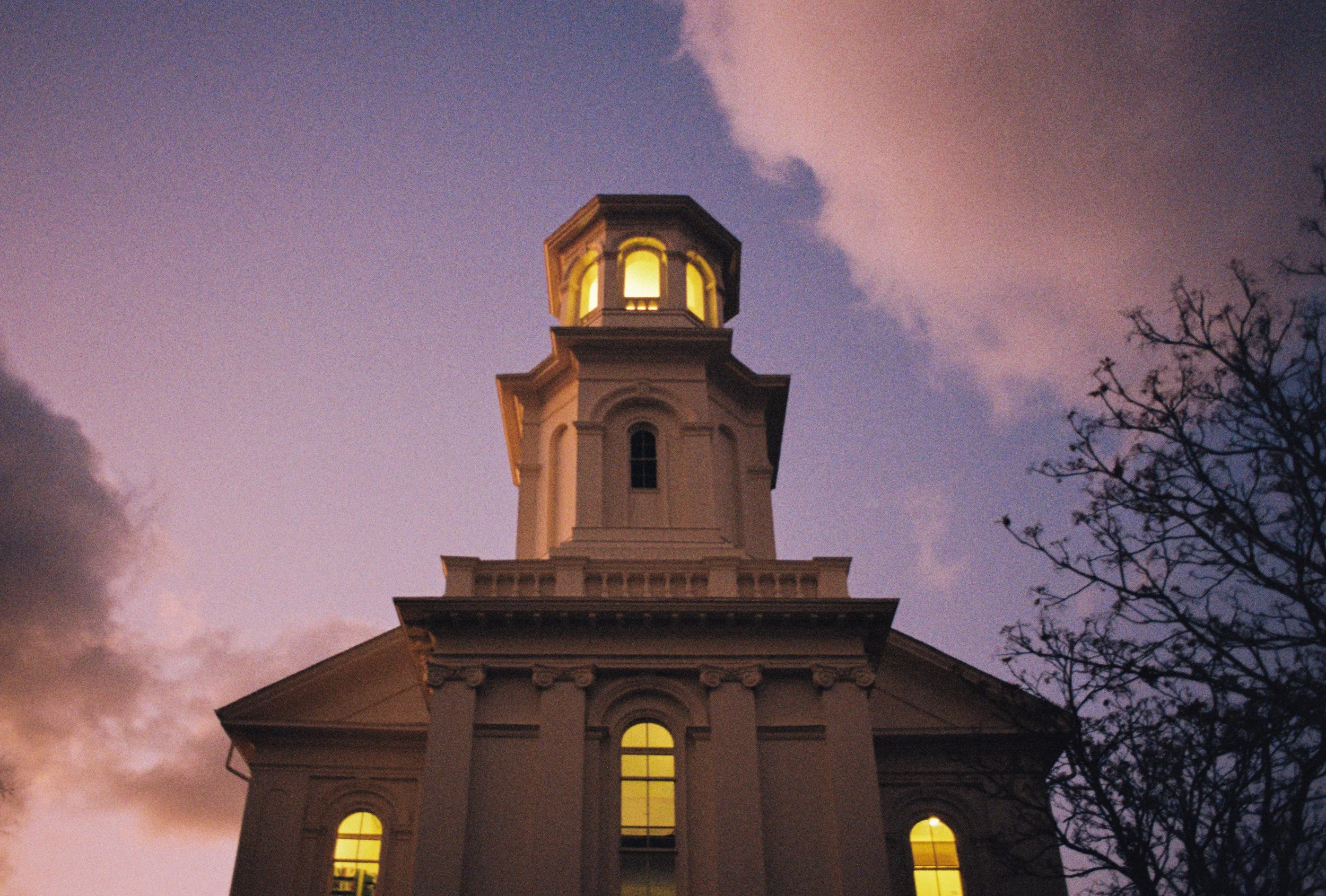 A tall, historic church building with a lit bell tower at dusk. The sky is a gradient of purple and pink, with clouds. A leafless tree is visible on the right.