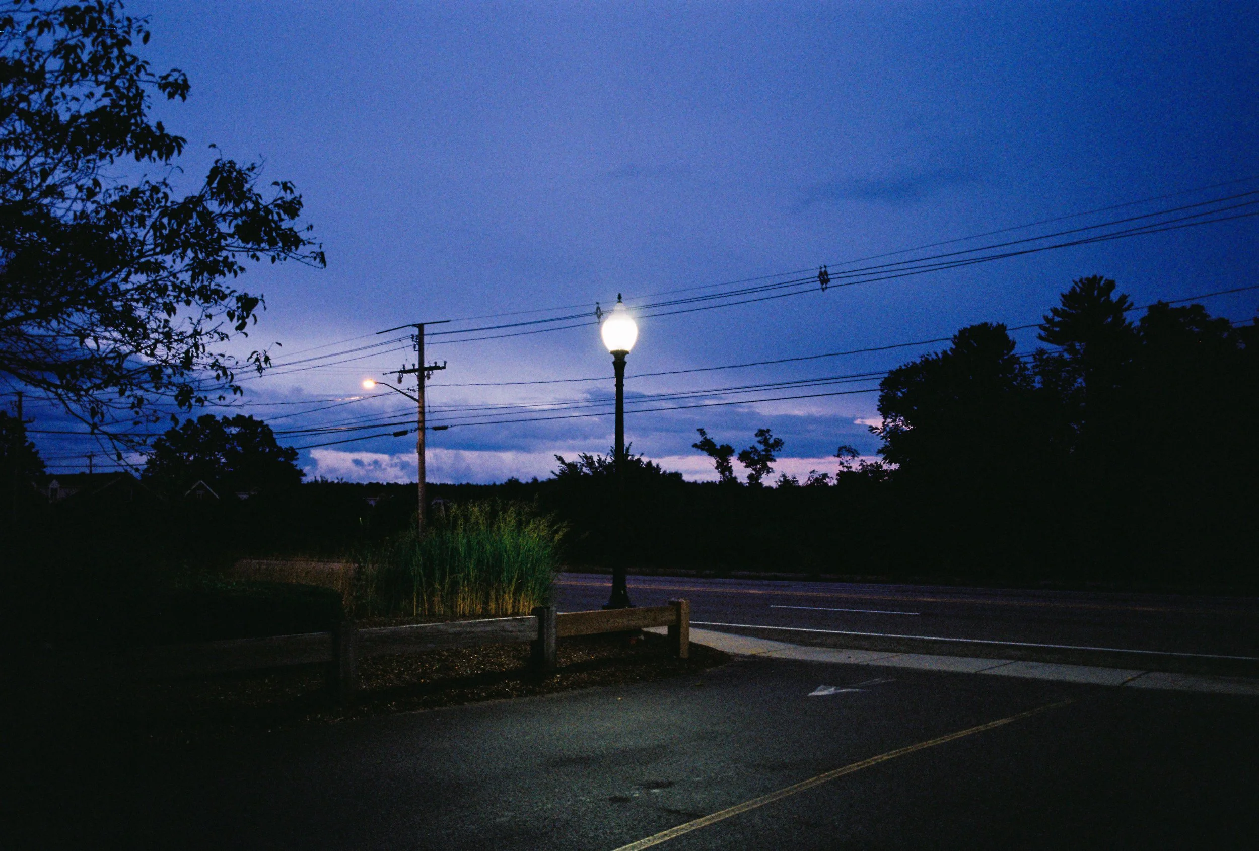 A street corner at dusk with a lit streetlamp, power lines, trees, and a darkening sky.